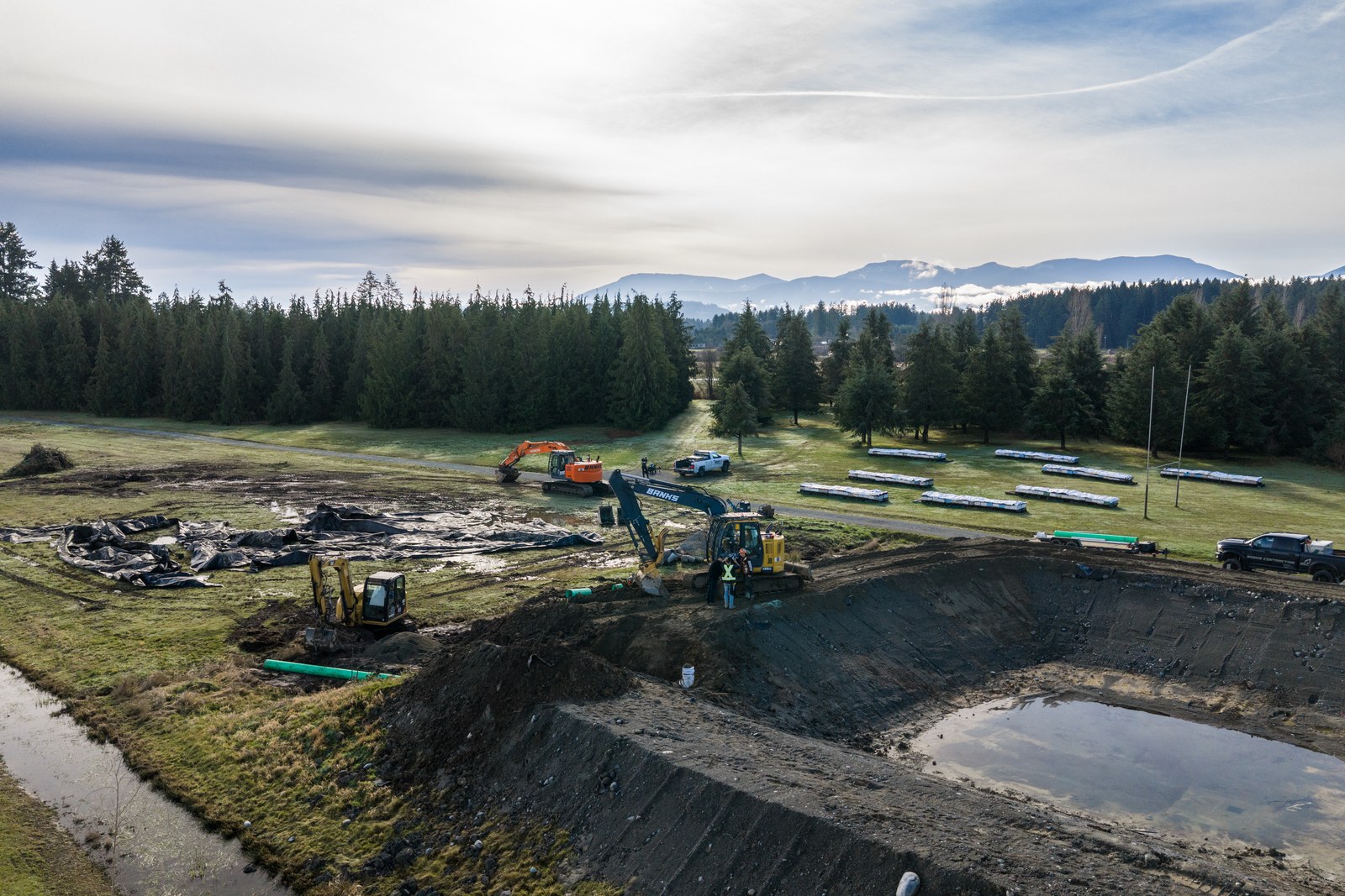 An aerial view of excavators, trucks and green pipe around a large excavated area bordered by trees and distant mountains