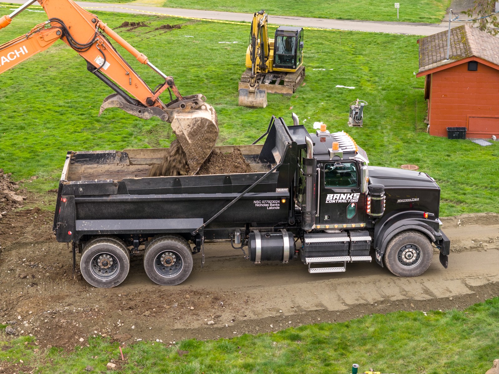 An elevated view of an orange excavator loading earth into a black Banks Contracting dump truck
