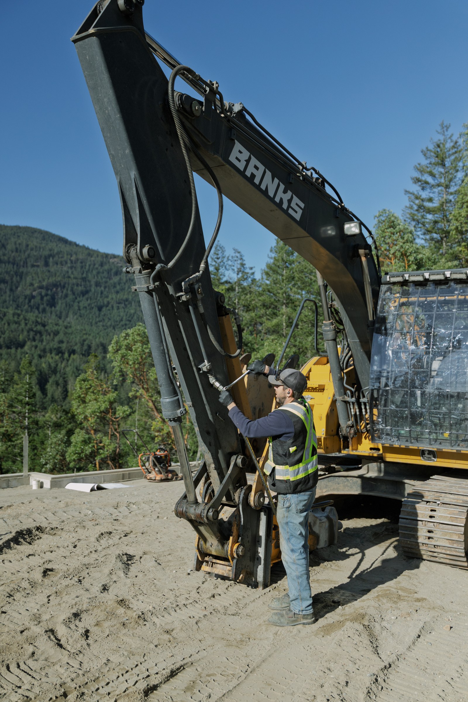 A worker in a reflective vest reaches up to an excavator attachment at a forested hillside site