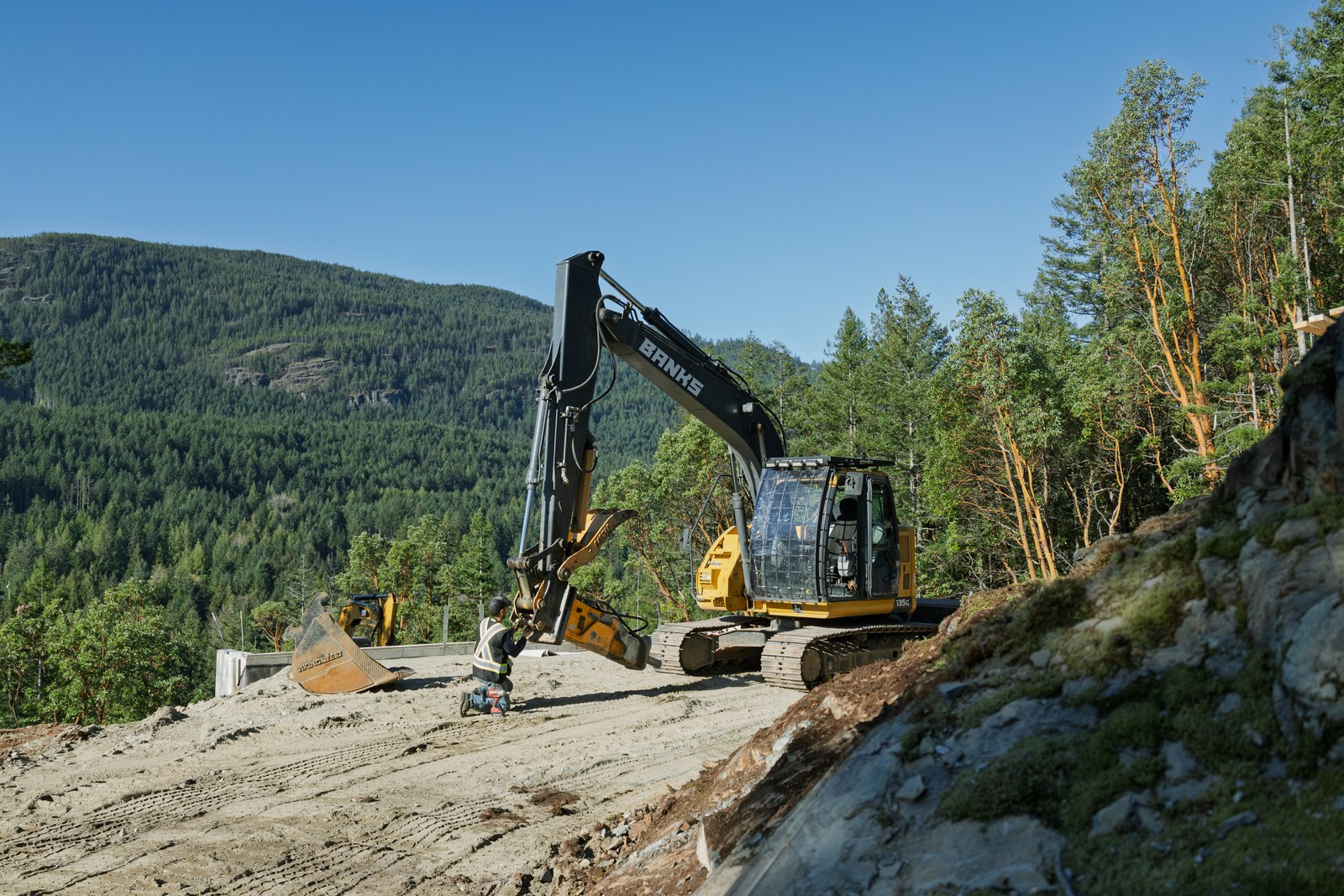 A worker kneels beside a Banks excavator at a hillside construction site surrounded by forest