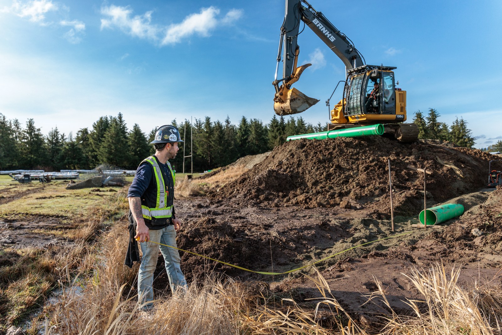 A worker in a reflective vest holds a measuring tape beside an excavator and green pipe at an earthworks site