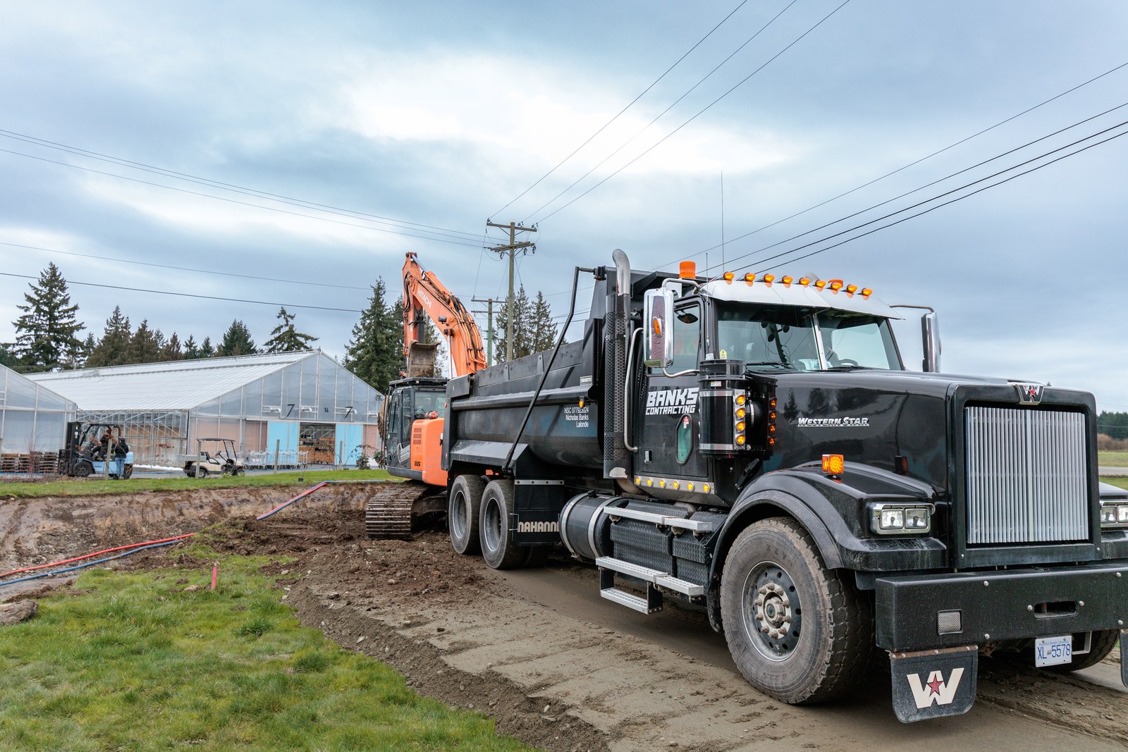 A black Banks Contracting dump truck stands beside an orange excavator and a greenhouse at an earthworks site
