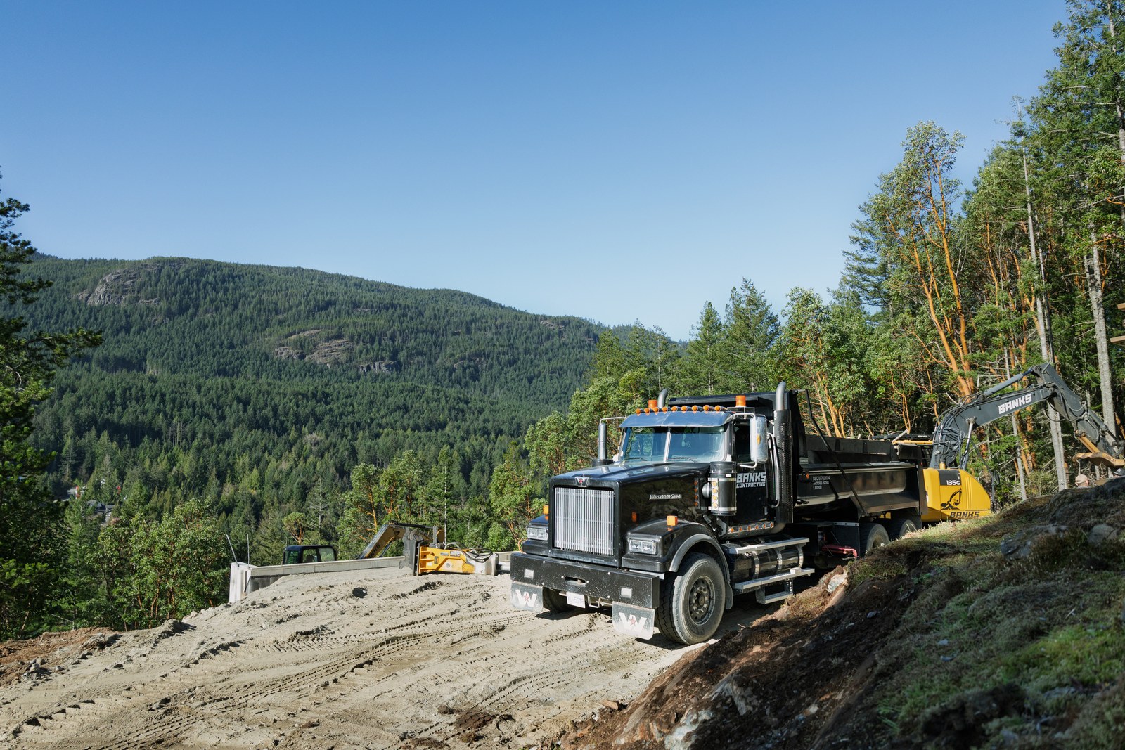 A black Banks Contracting dump truck and yellow excavator stand on a hillside road above a forested valley