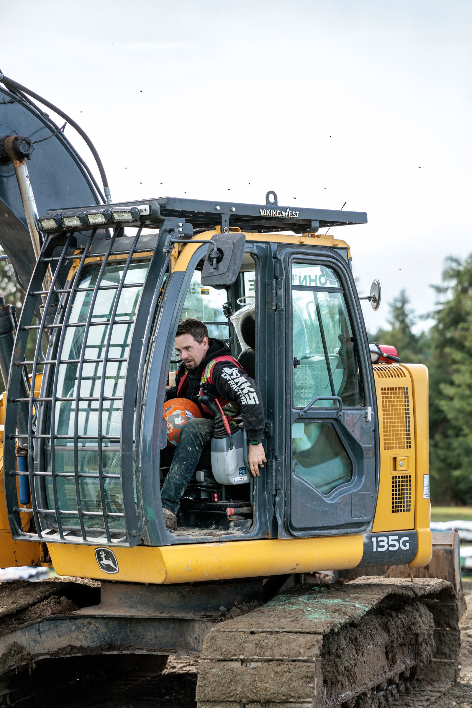 A Banks Contracting operator sits in the open cab of a yellow excavator holding an orange hard hat