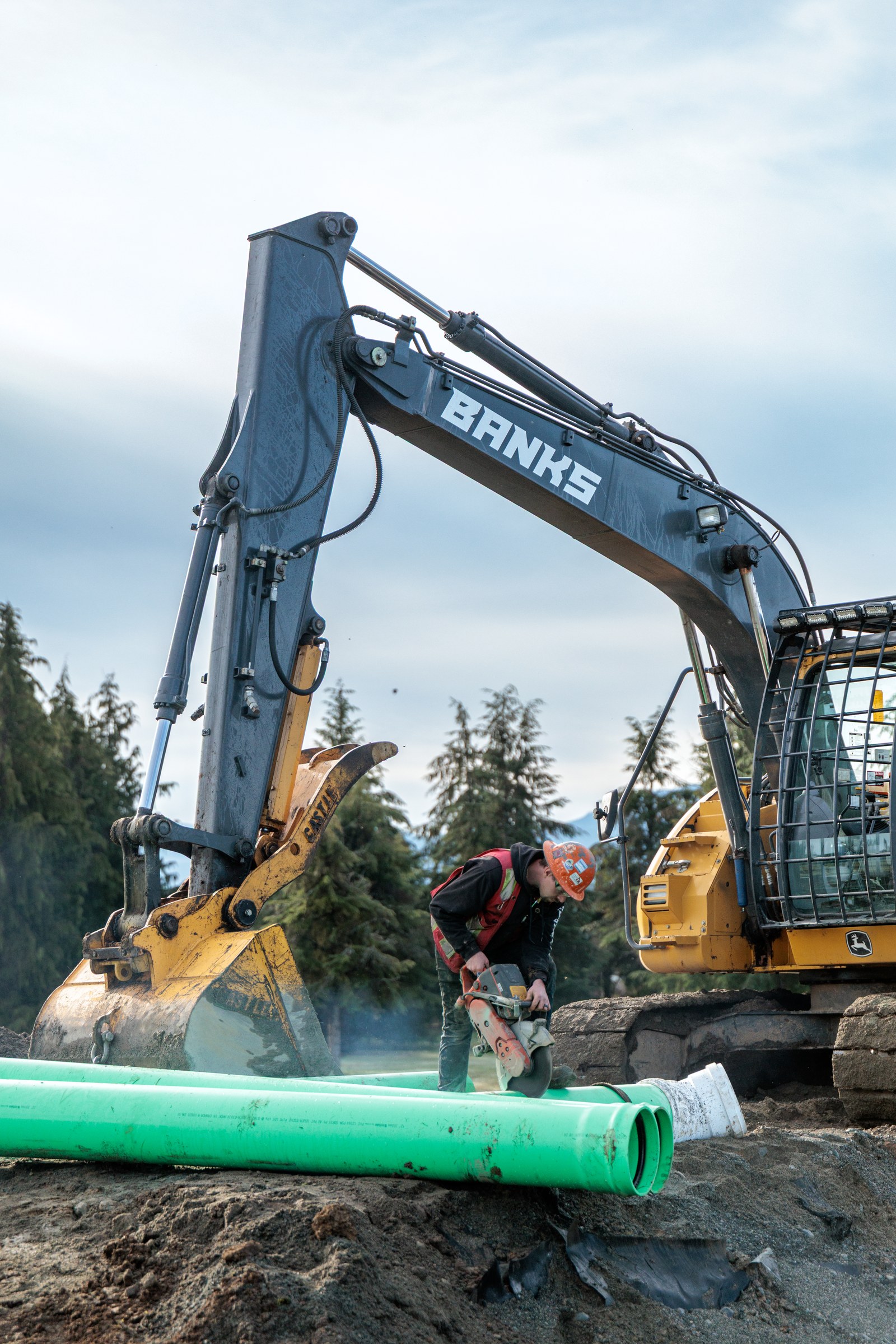 A worker in an orange hard hat uses a cut-off saw beside green pipe and a Banks excavator