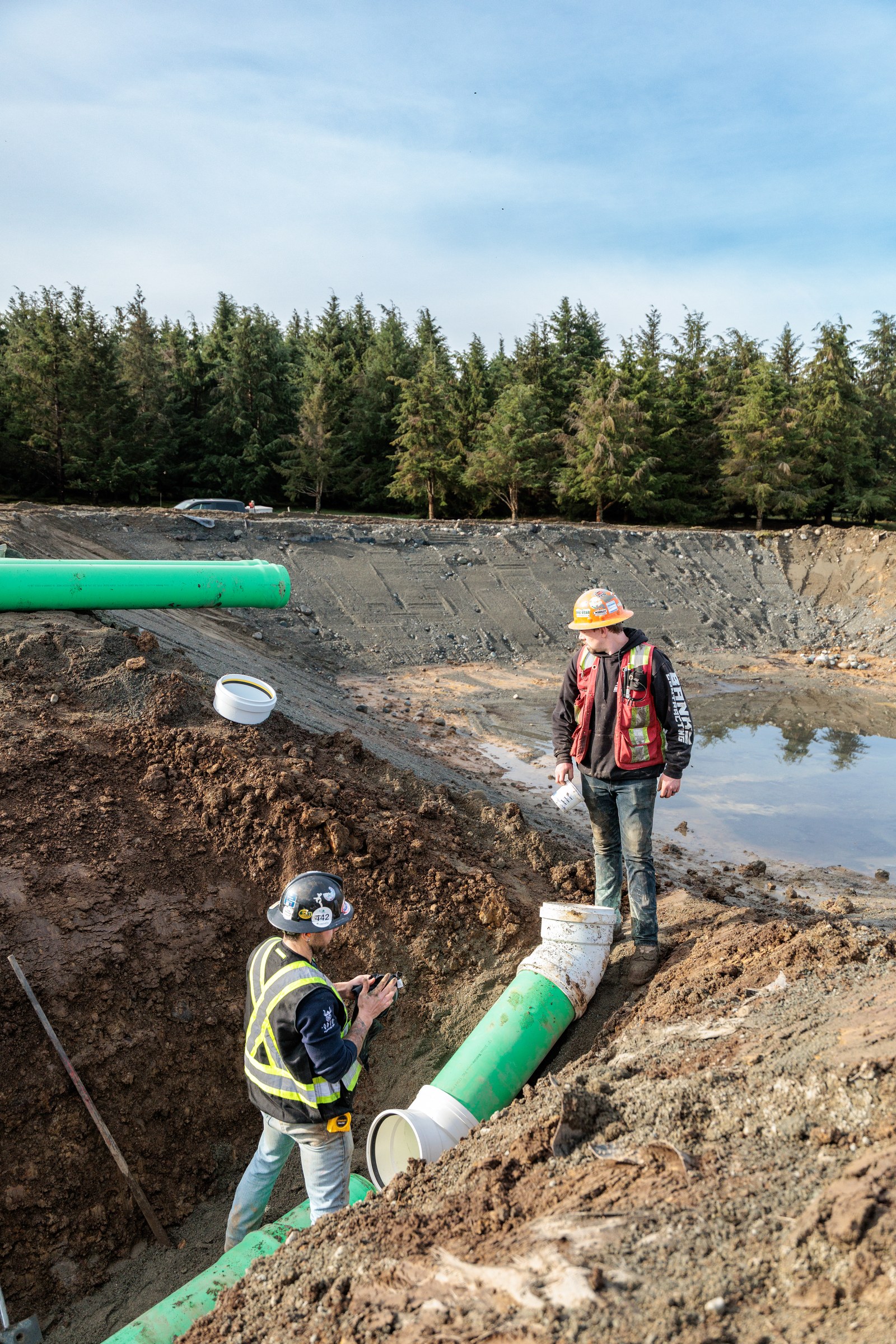 Two workers in hard hats stand at different levels beside green pipe in an excavated trench
