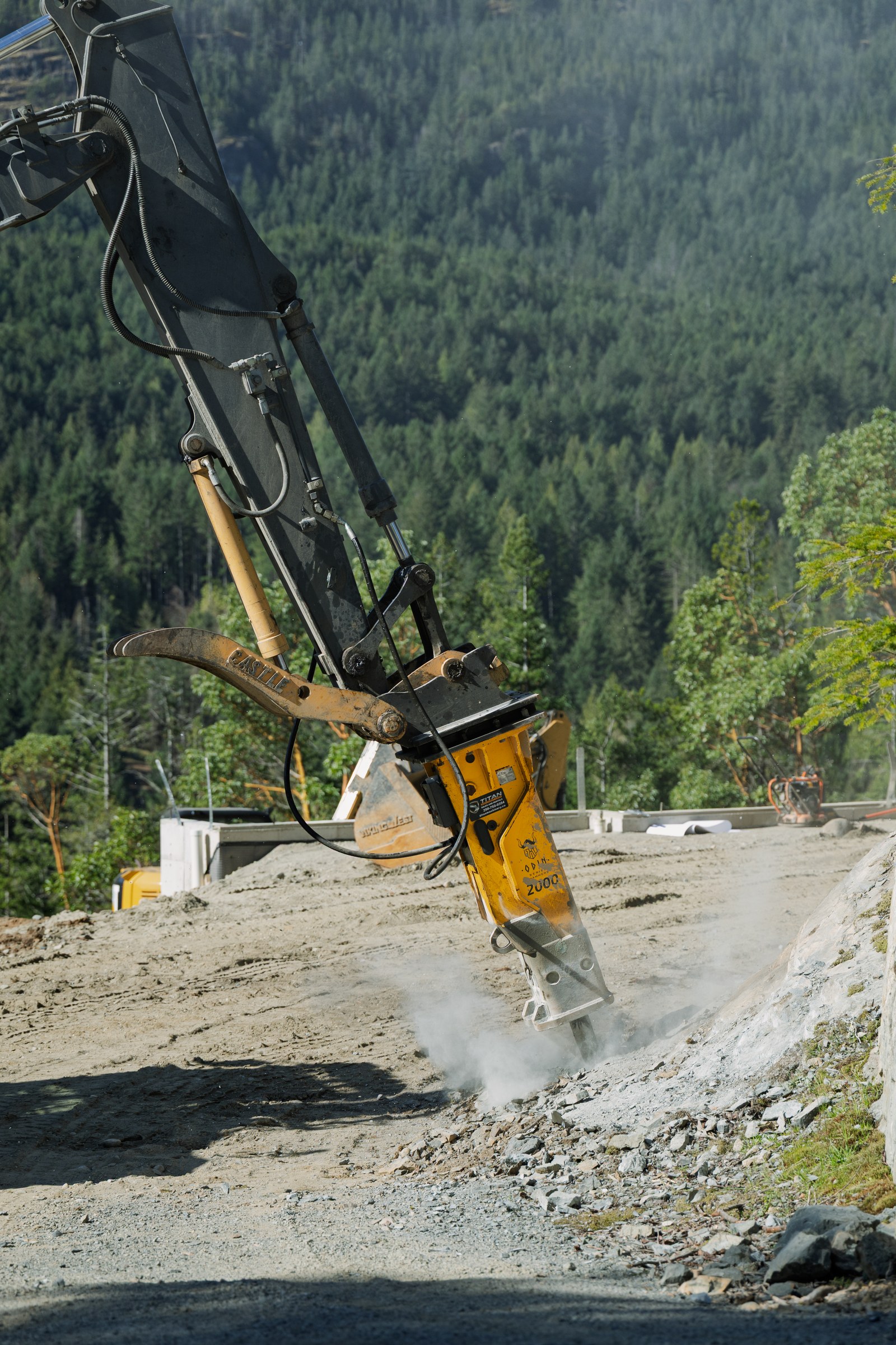 A yellow hydraulic breaker attached to an excavator breaks exposed rock with dust rising in front of a forested mountain