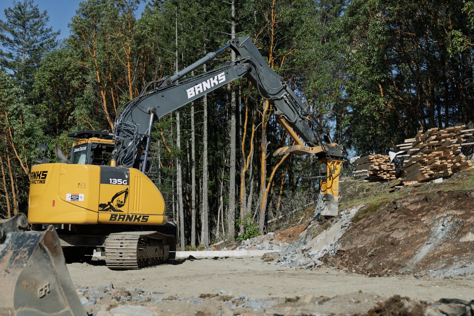 A yellow Banks excavator uses a hydraulic breaker on exposed rock beside forest and stacked lumber