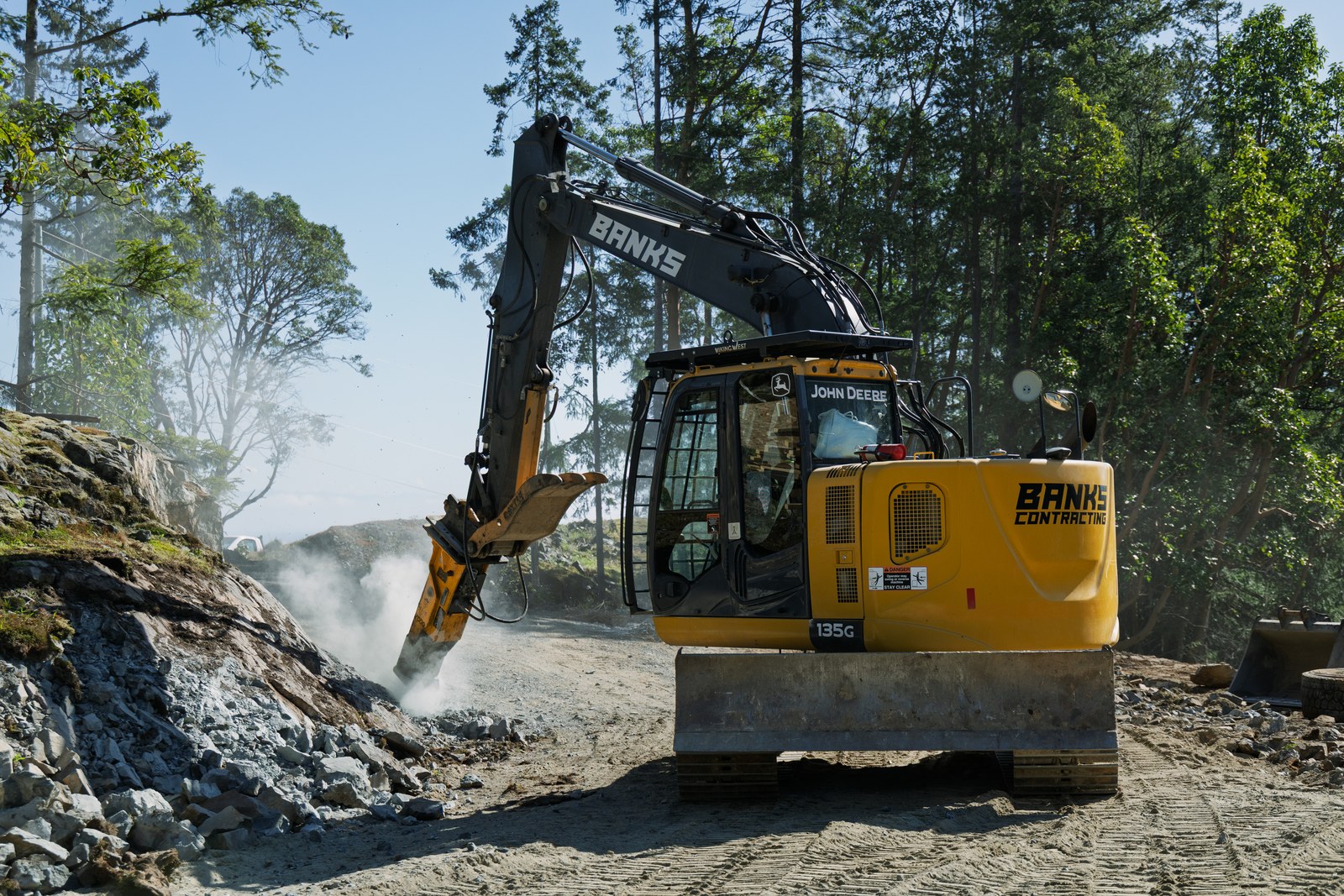 A Banks Contracting excavator breaks rock as backlit dust rises among trees