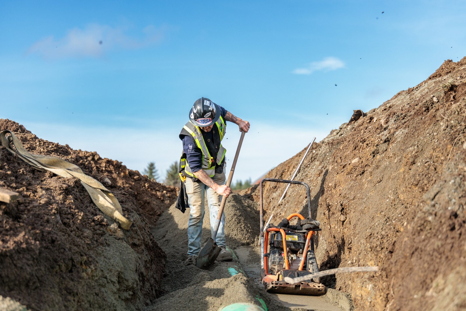 A worker in a black hard hat shovels soil beside a plate compactor in a trench under a blue sky