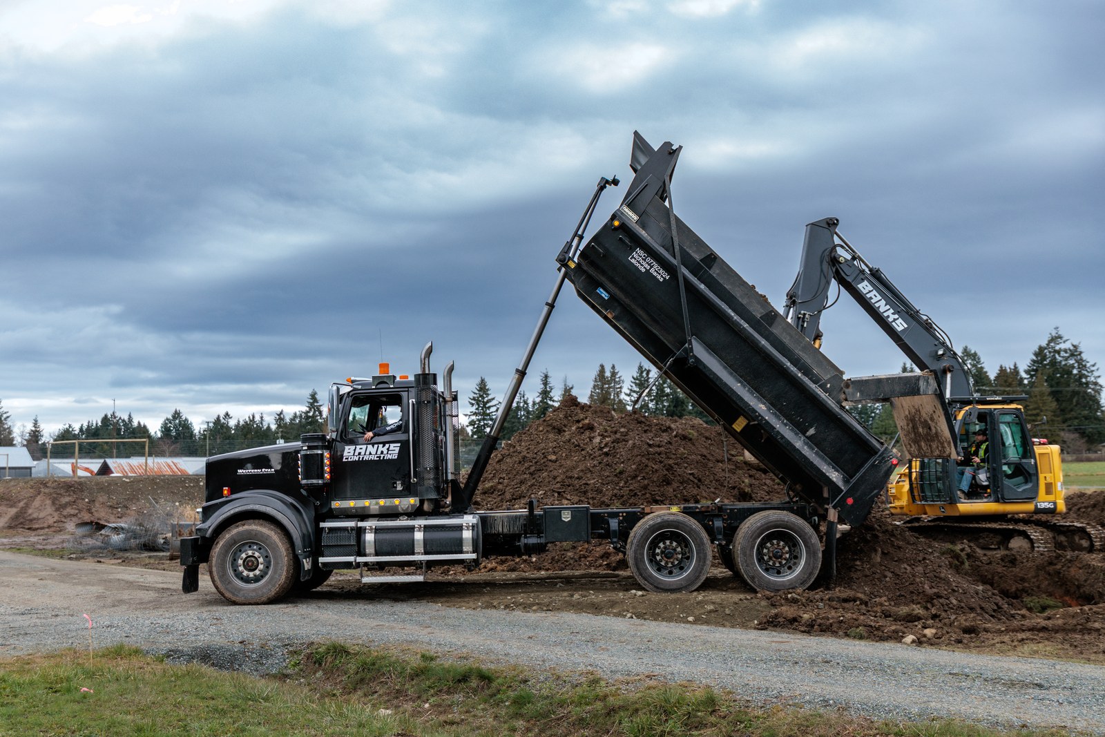A black Banks Contracting dump truck tips earth beside a yellow excavator under a cloudy sky