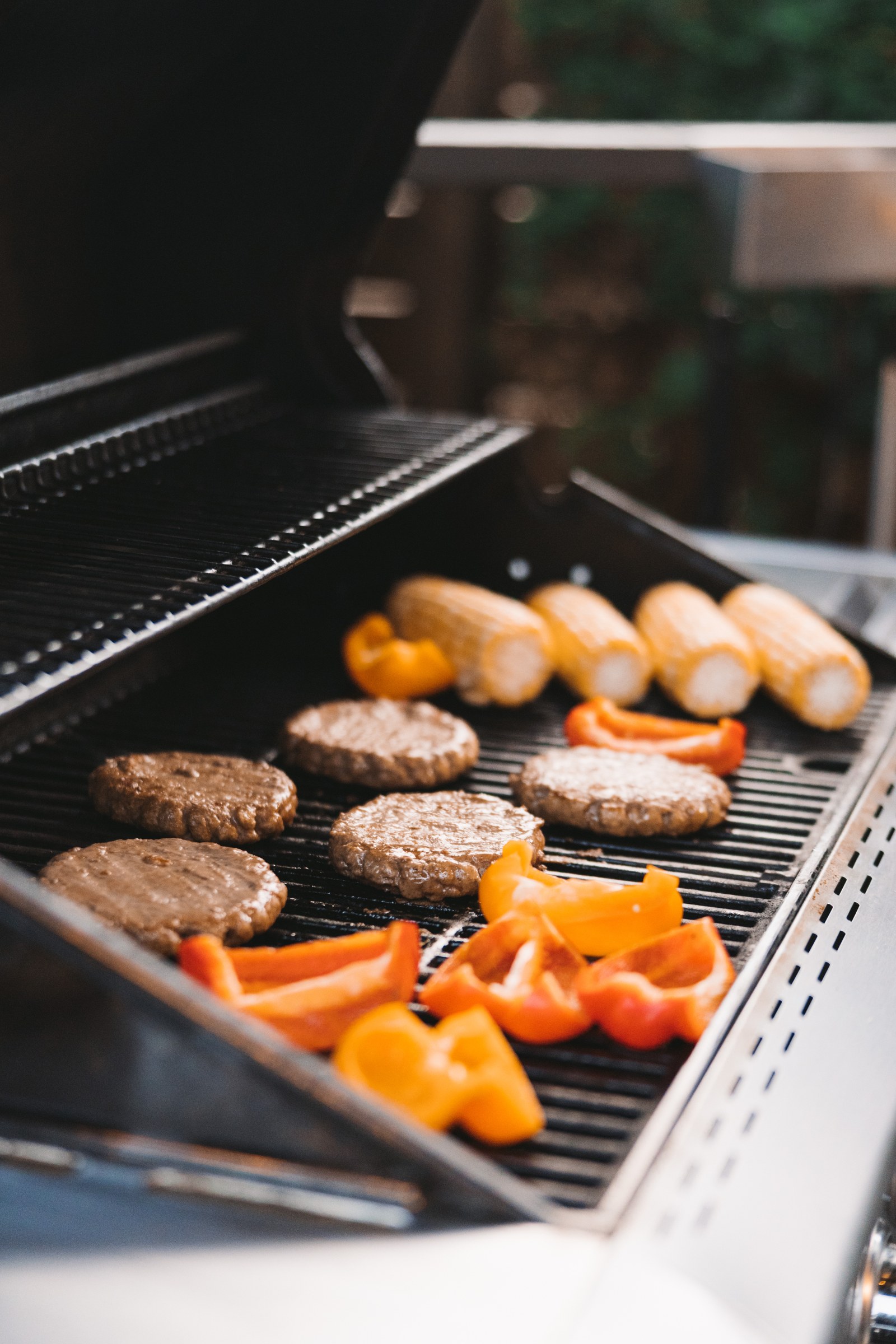 Burger patties, corn and sliced peppers cook on an open barbecue grill