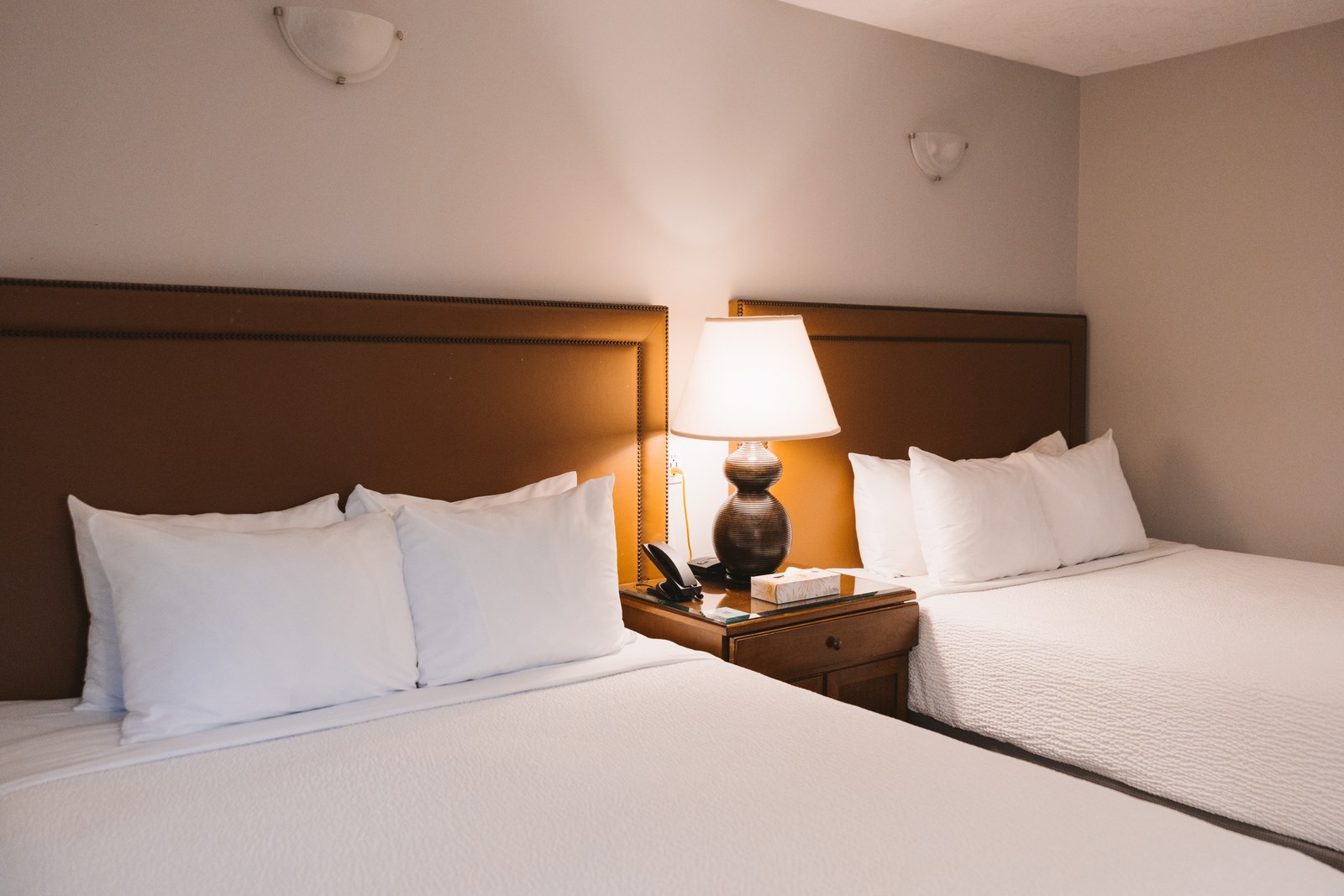 Two neatly made hotel beds with white bedding sit beside a shared bedside table and warm lamp