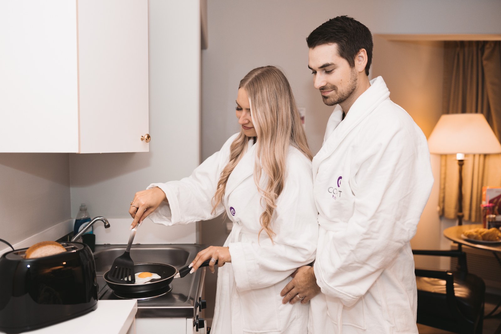 Two guests in Coast robes cook breakfast together in a compact kitchen