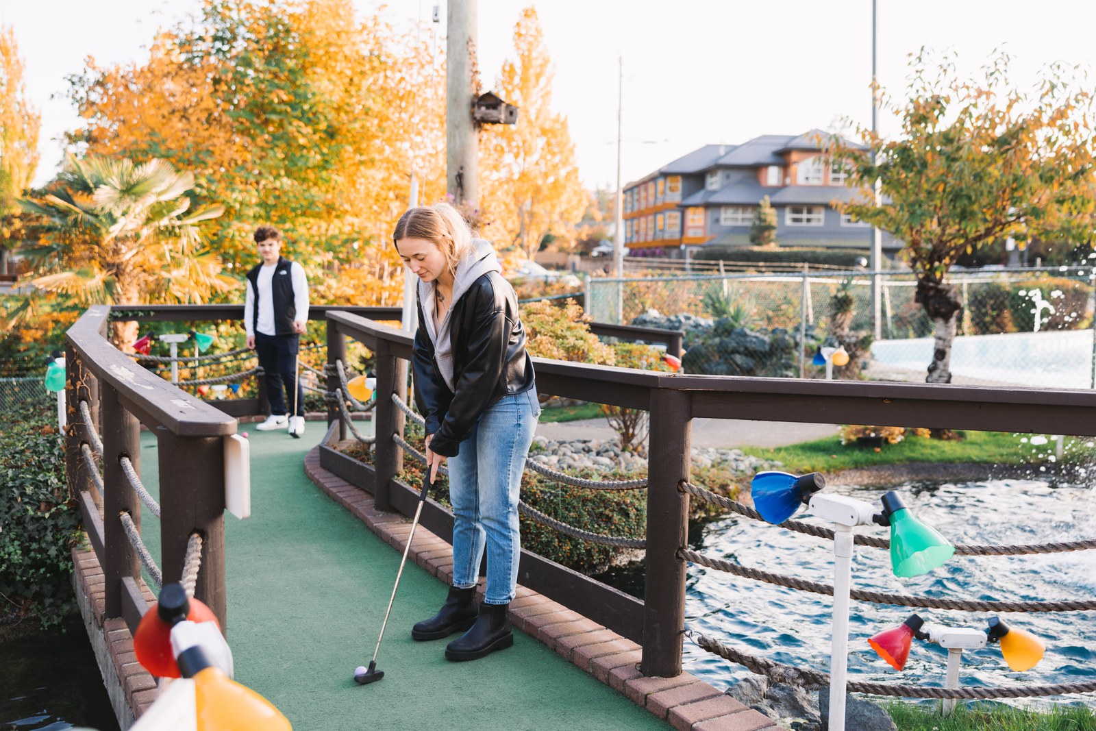 Two guests play mini golf beside a pond with autumn trees in the background