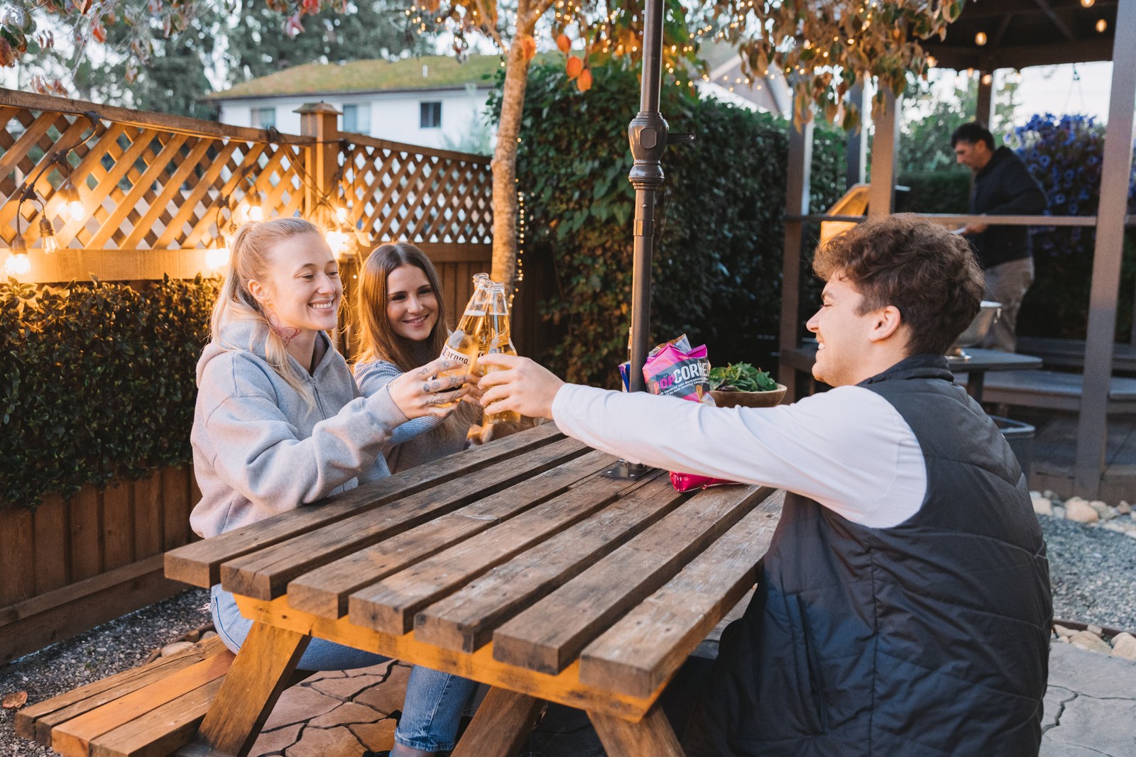 Three guests clink bottles at a wooden patio table beneath string lights