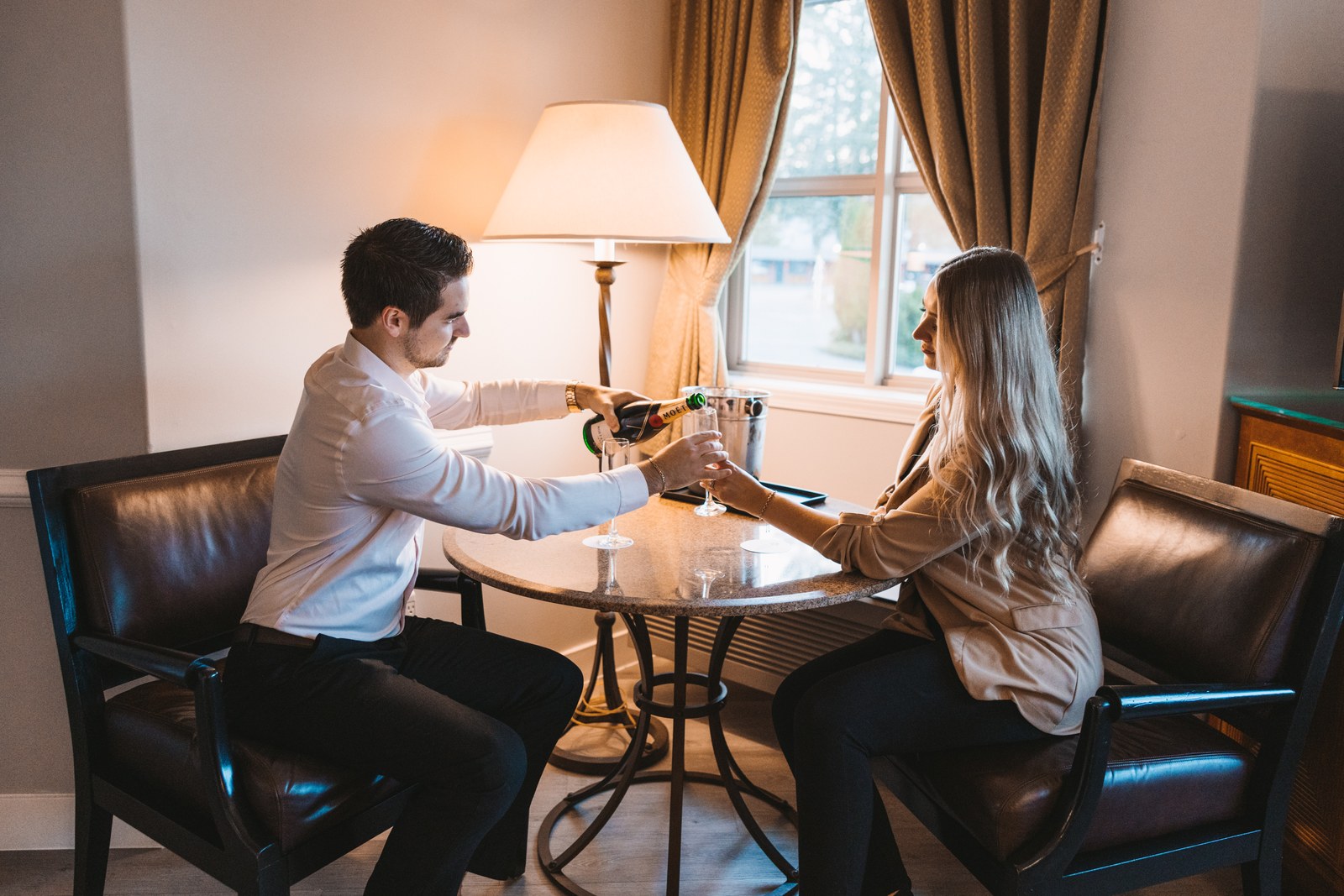 Two guests sit at a table beside a hotel window while champagne is poured into their glasses