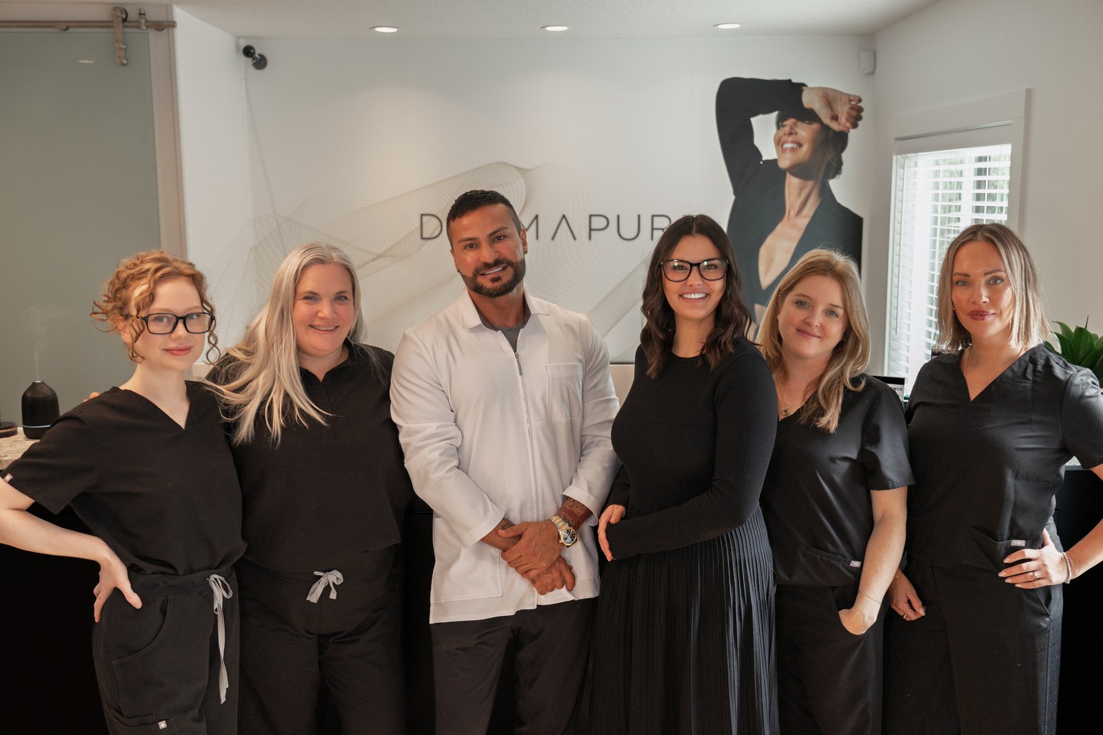 Six clinic team members posing together in front of a Dermapure reception wall