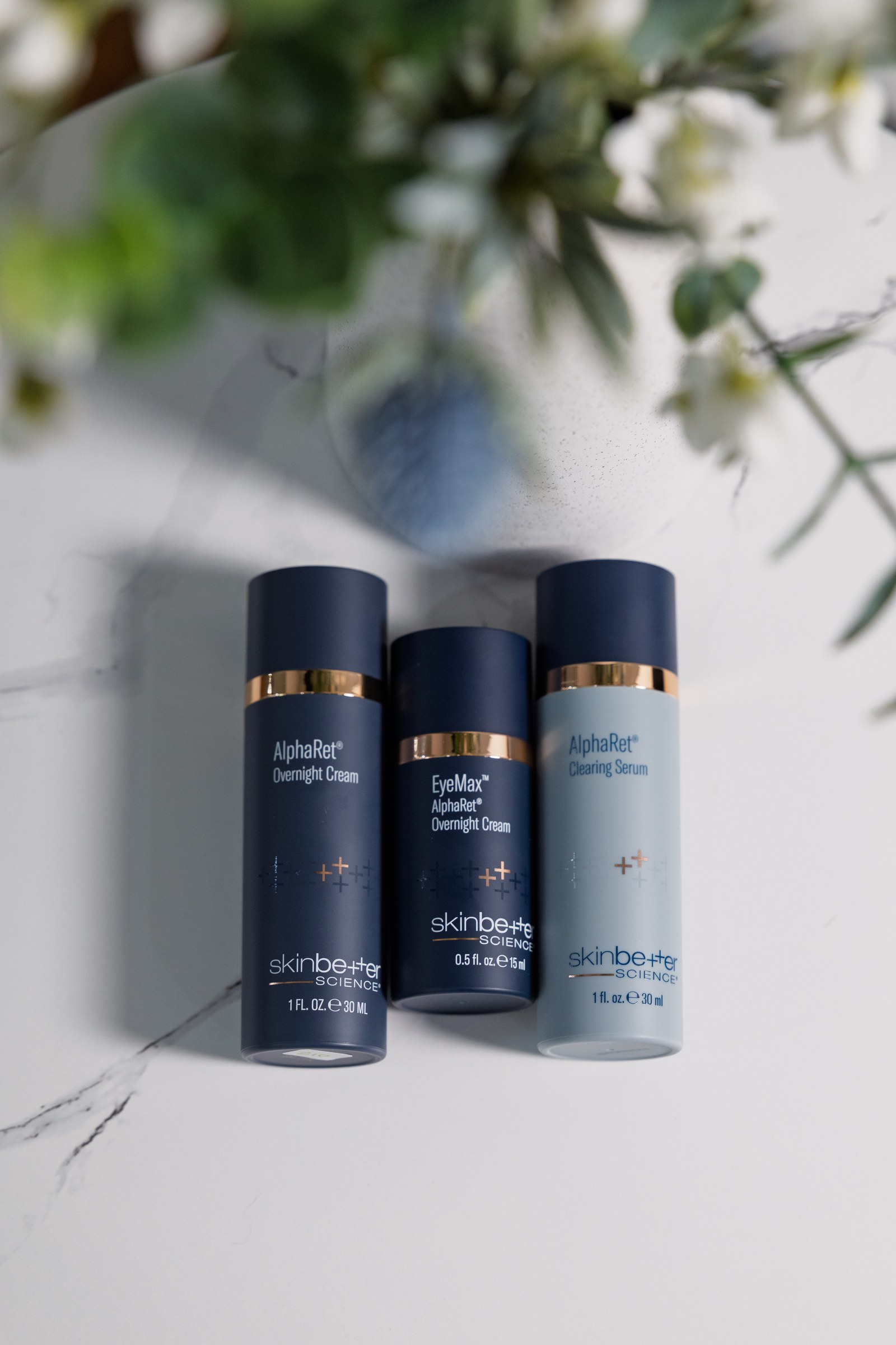 Three blue and grey skincare bottles arranged on a pale surface beneath softly blurred flowers