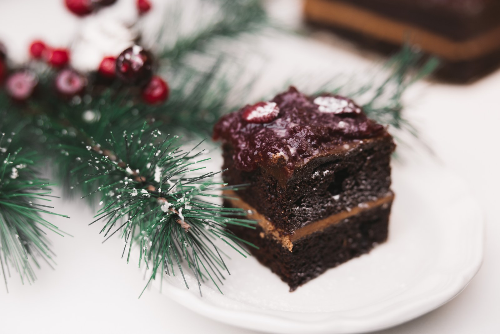 Layered chocolate dessert with cranberry topping on a white plate beside evergreen branches