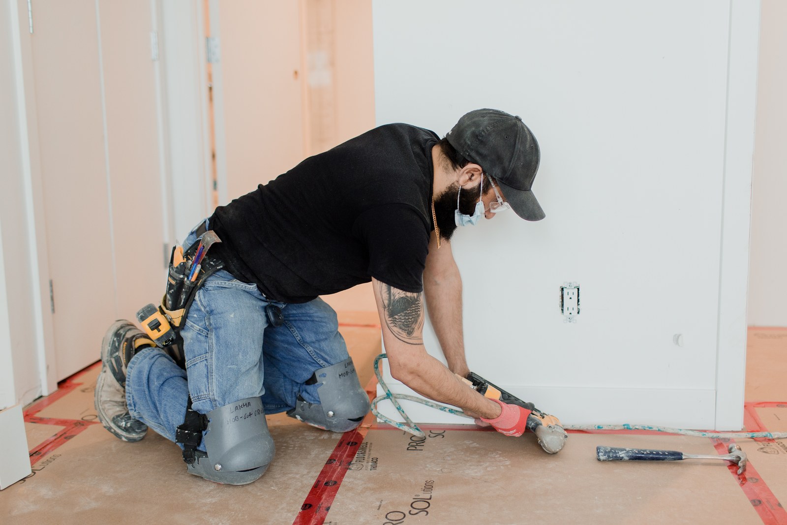 A carpenter kneels beside a wall and uses a nail gun to install white baseboard trim