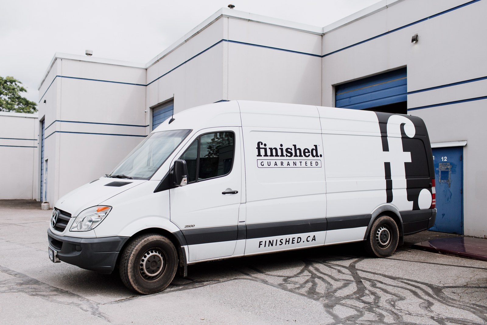 A white Finished branded work van is parked outside a workshop with blue doors