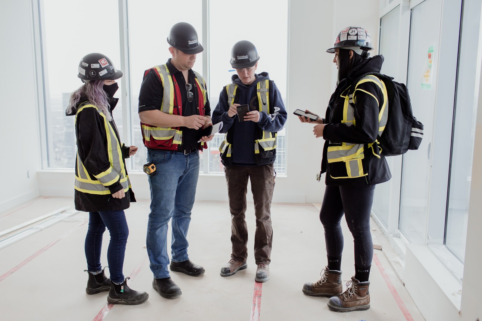 Four construction crew members in hard hats discuss work beside bright floor-to-ceiling windows