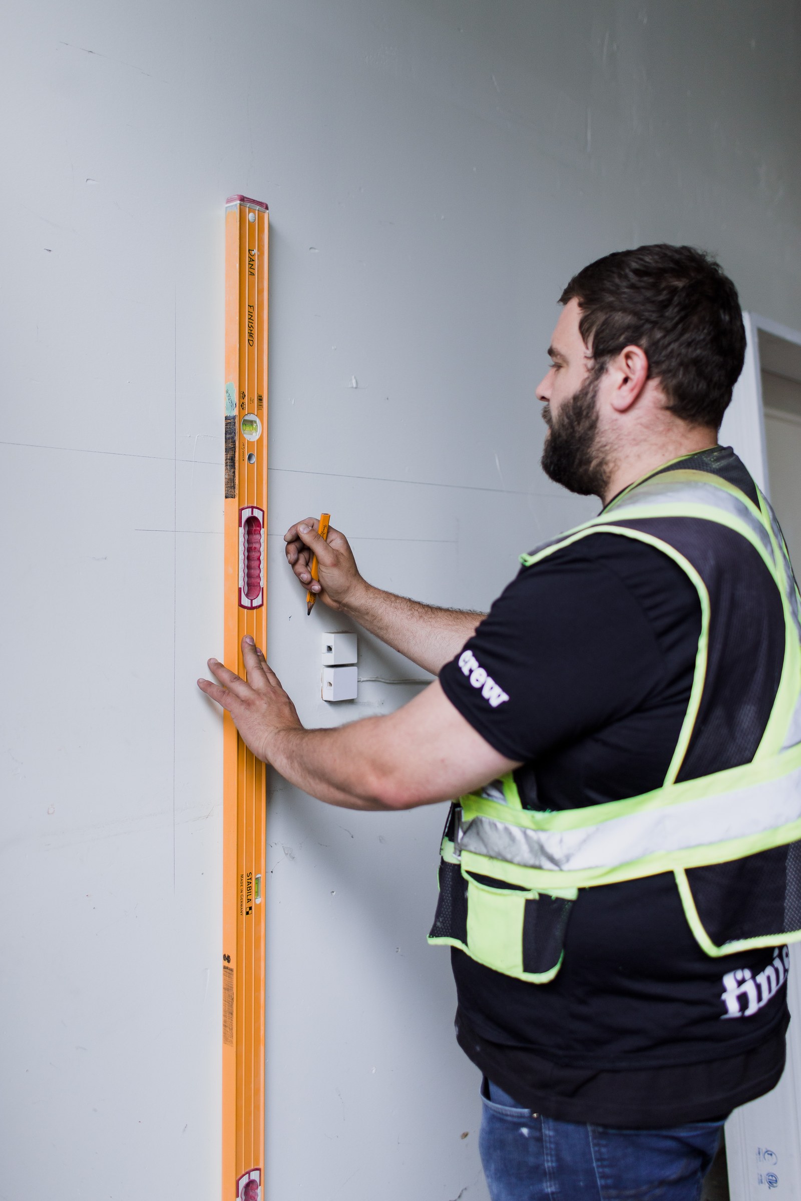 A carpenter holds a tall orange spirit level against a wall and marks a line with a pencil