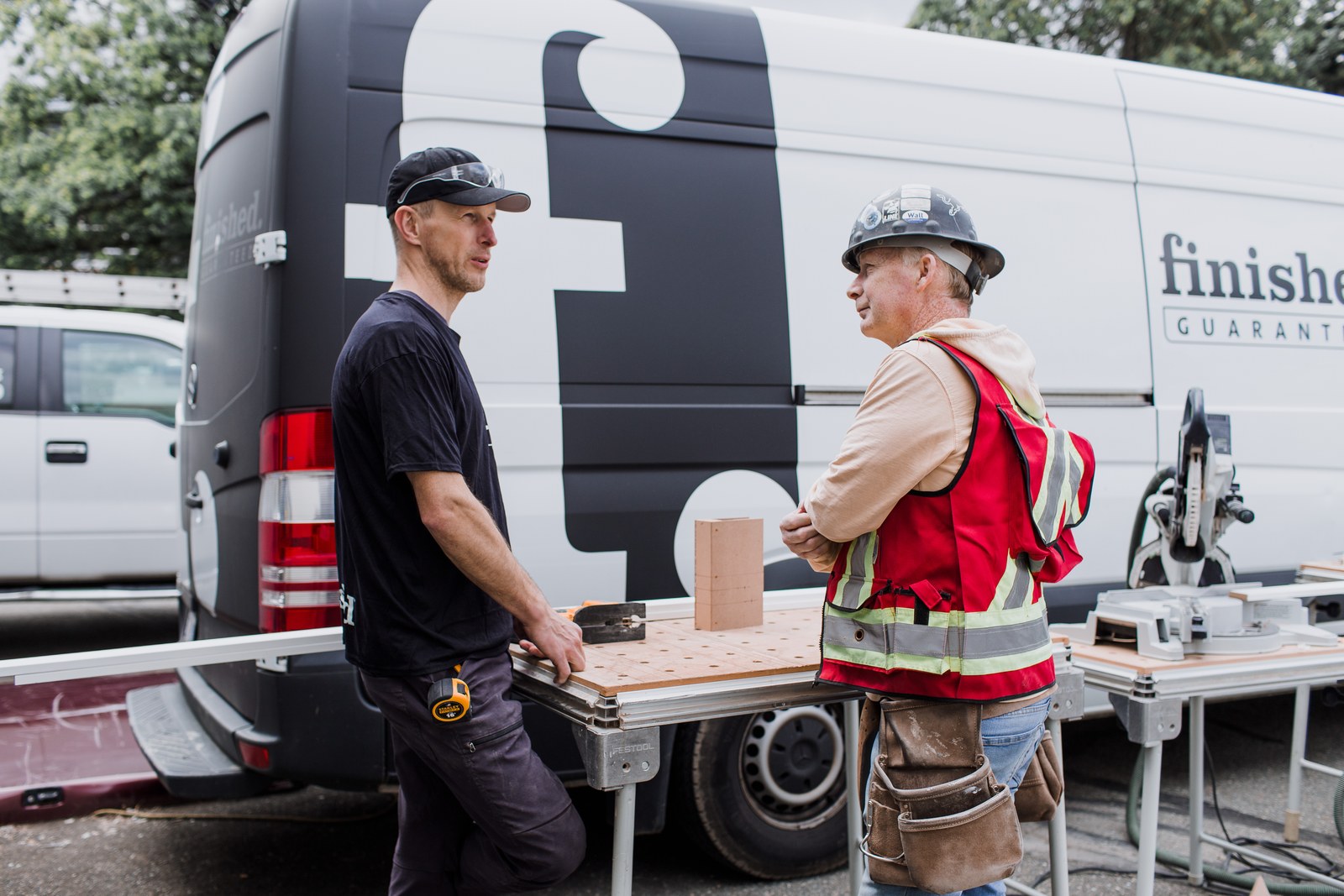 Two carpenters talk beside a workbench in front of a Finished branded van