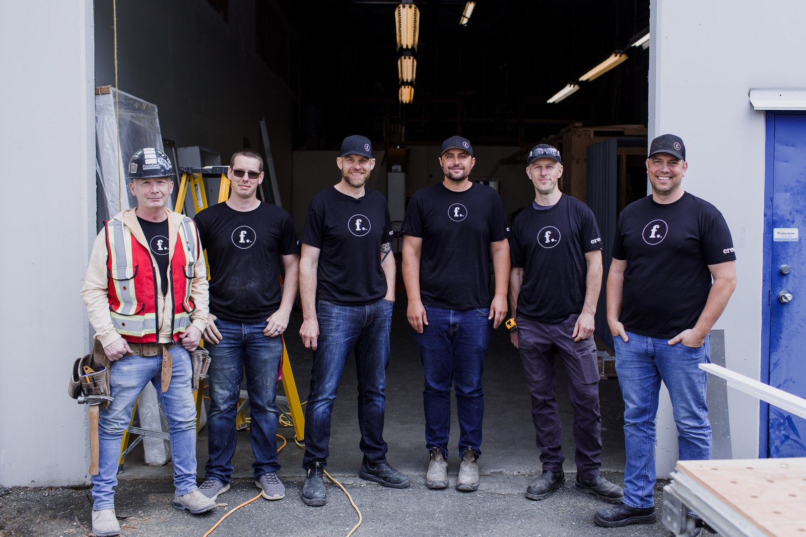 Six Finished team members in branded black shirts stand outside an open workshop
