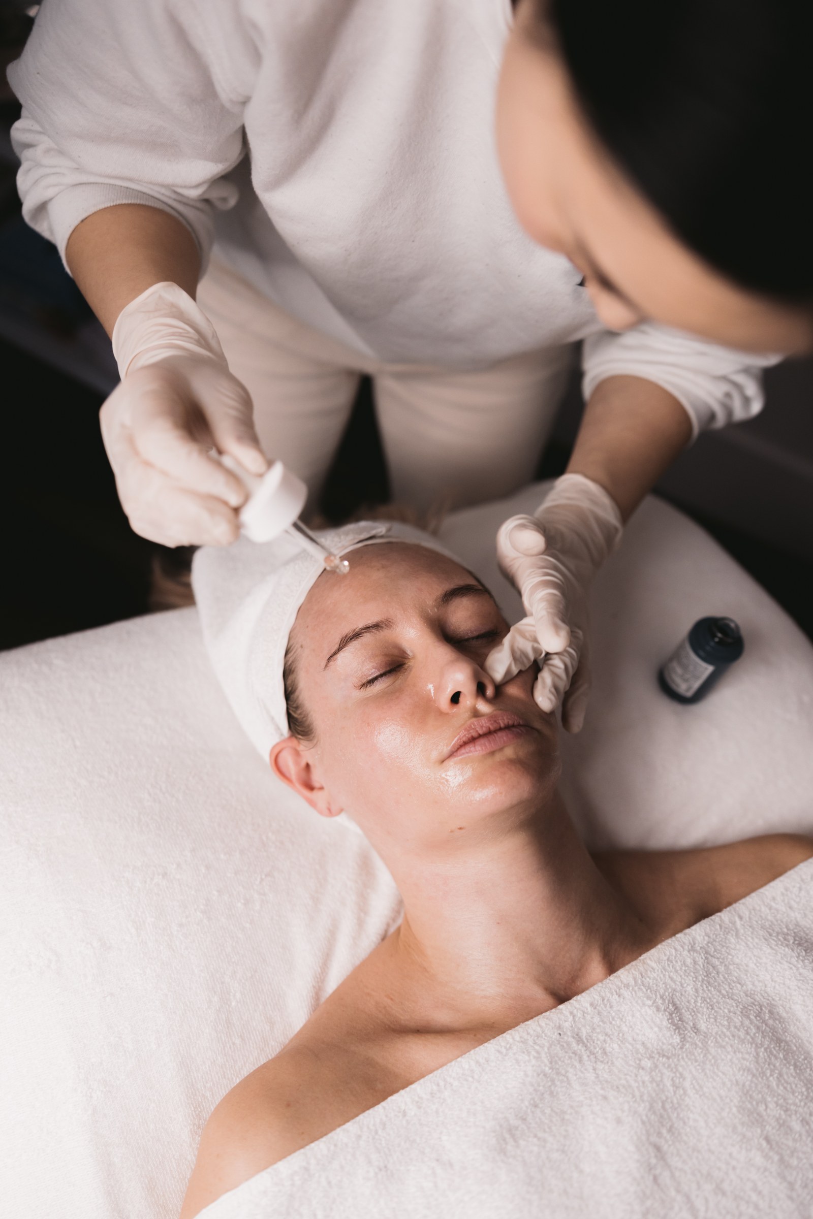 A gloved practitioner applies liquid from a dropper to a client’s forehead during a facial treatment