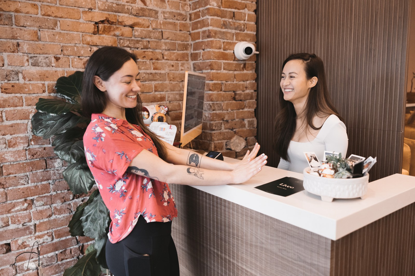 Two women smile across a white reception counter in a spa with an exposed brick wall