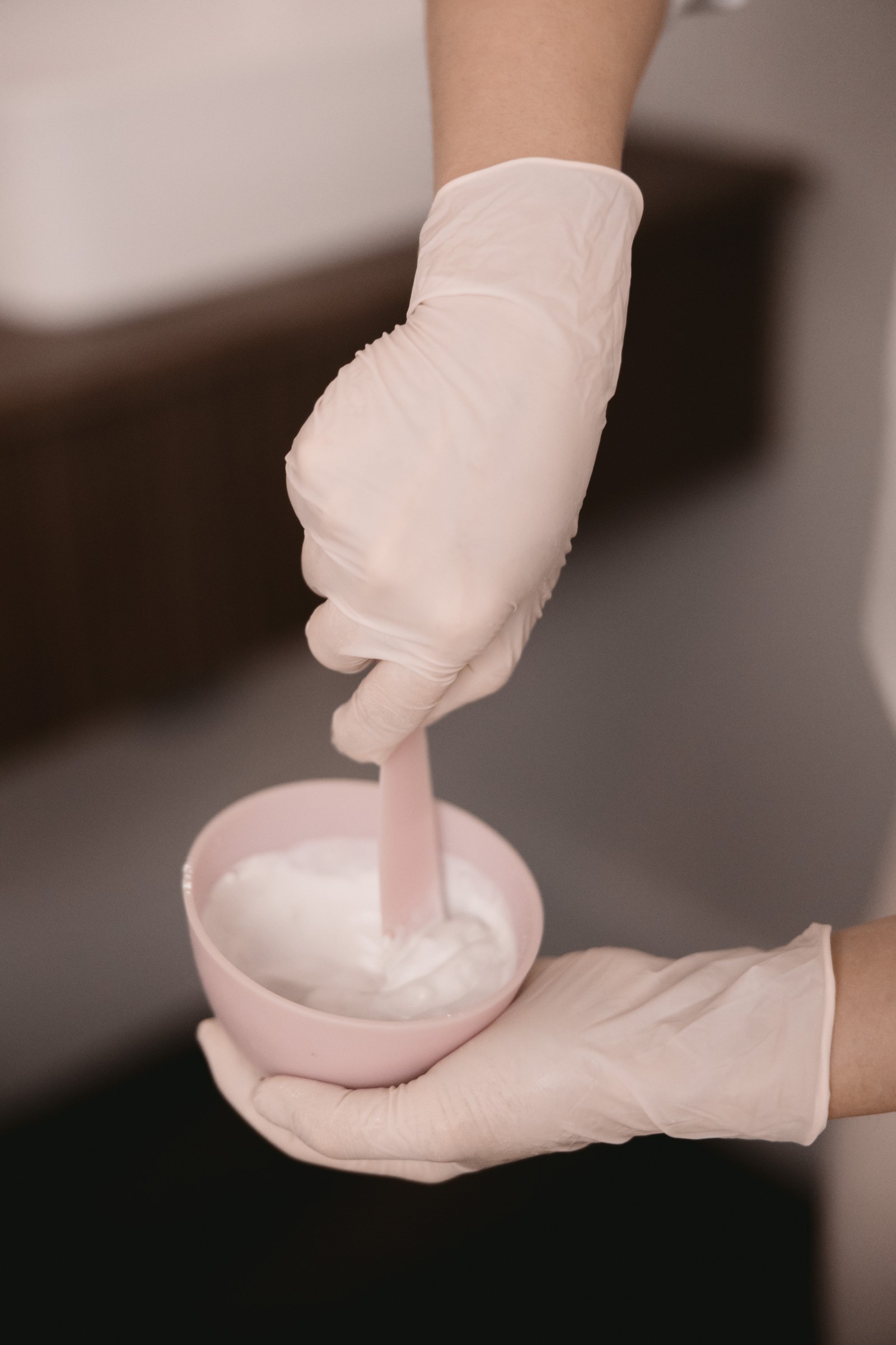 Gloved hands mix a white skincare treatment in a small pink bowl