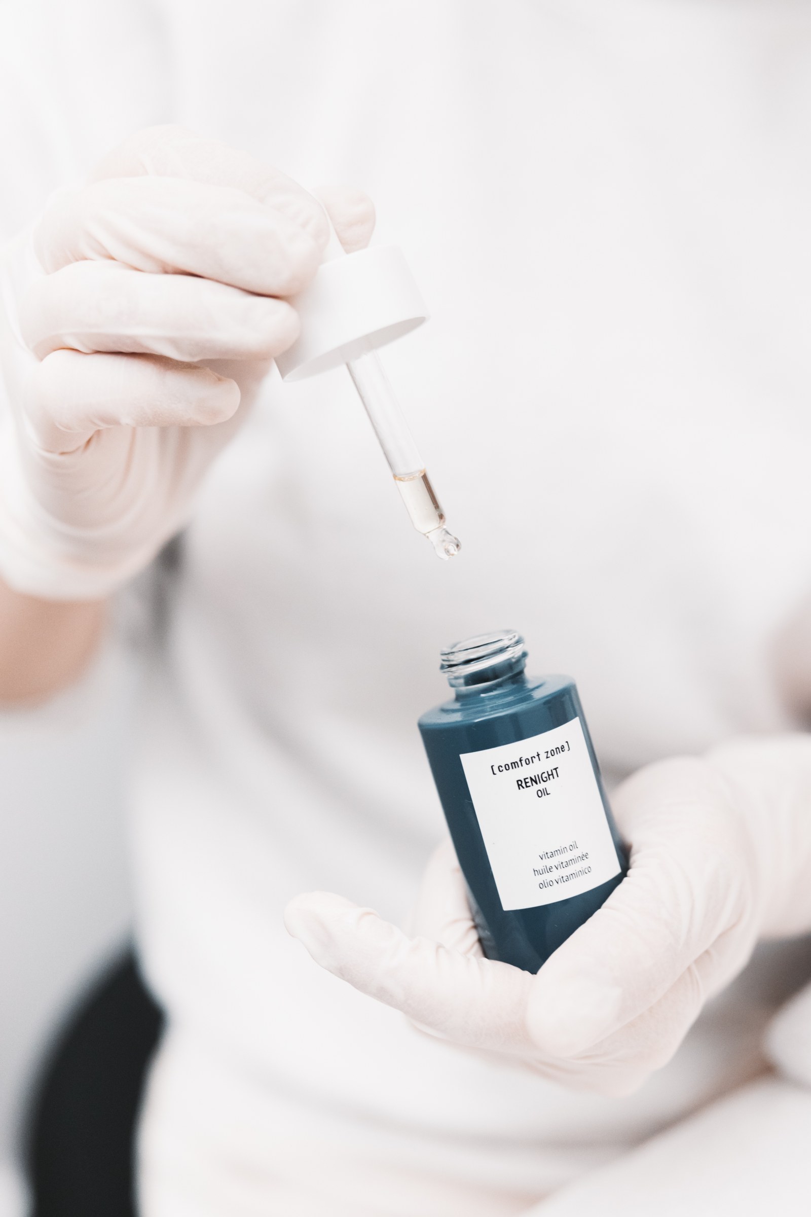 Gloved hands hold a blue skincare oil bottle and a glass dropper with a suspended droplet