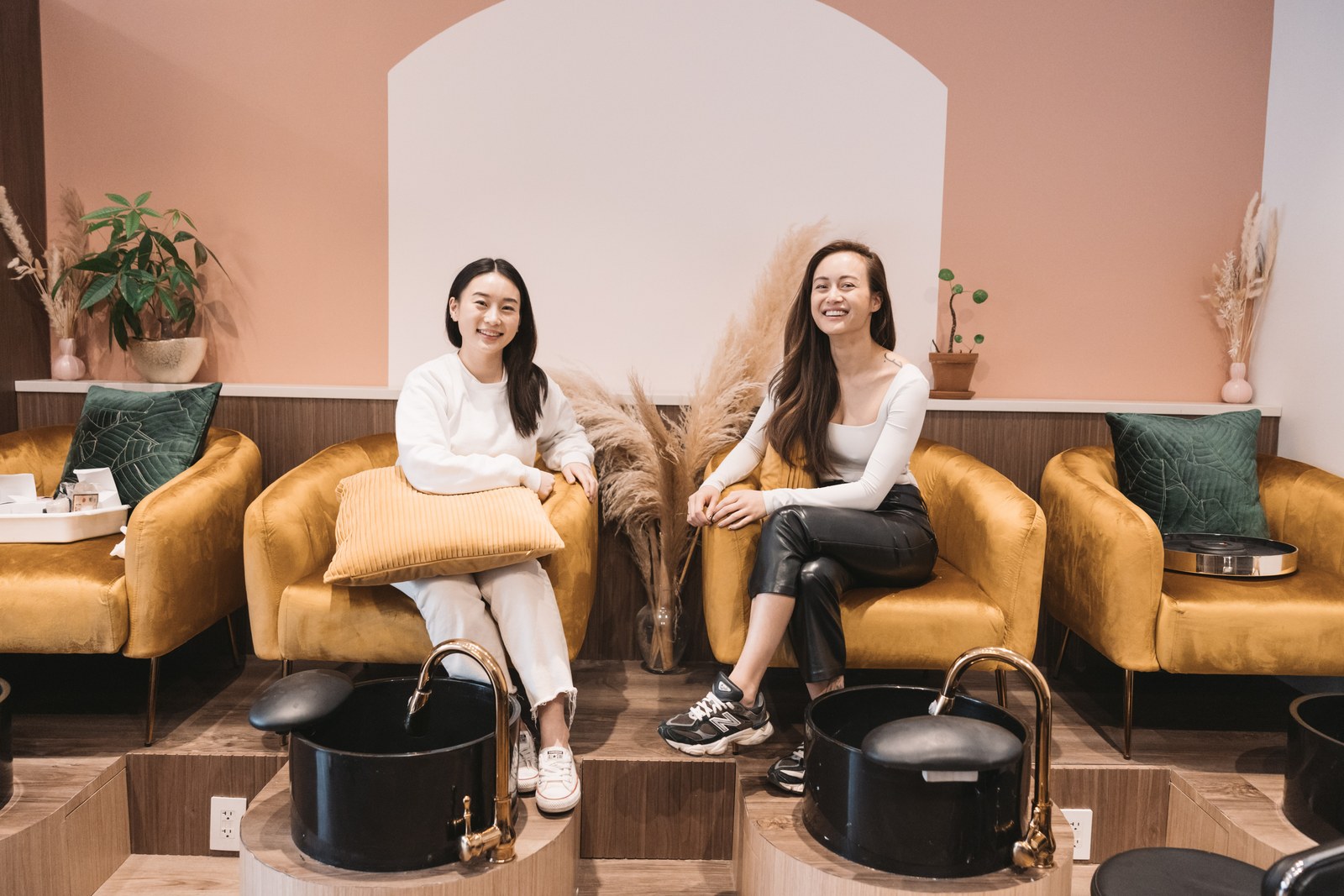Two women sit smiling on gold chairs beside pedicure basins in a pink and wood-toned spa interior