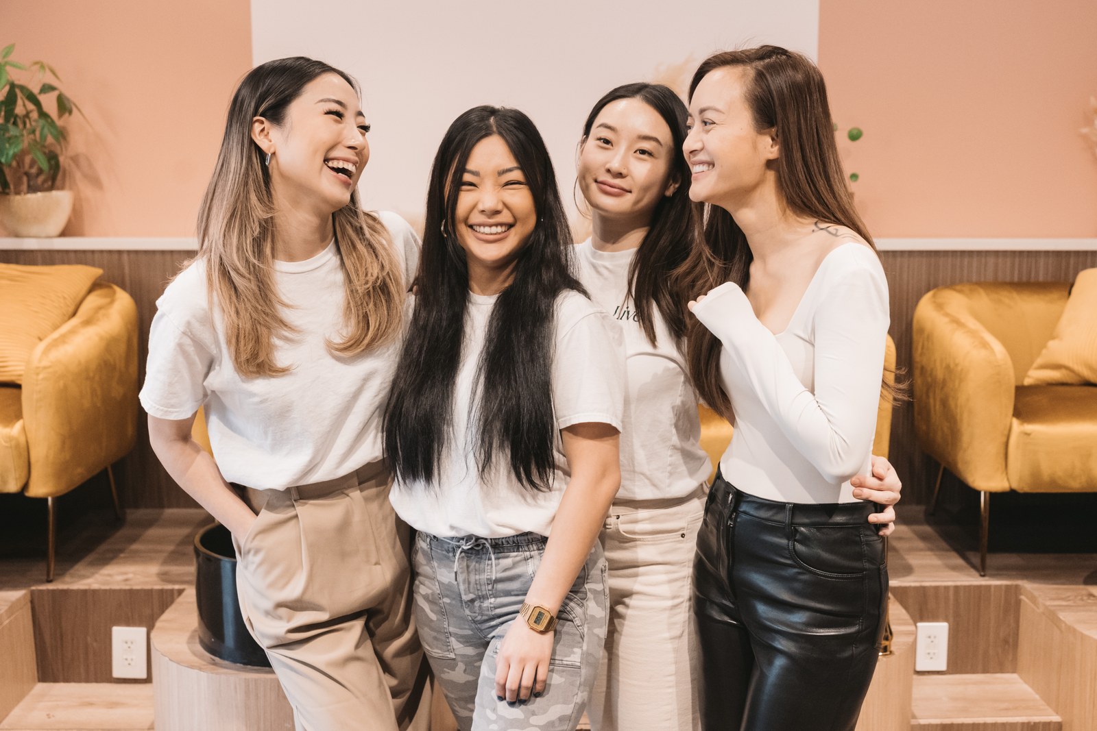 Four women smile together in a spa with gold chairs and pink walls