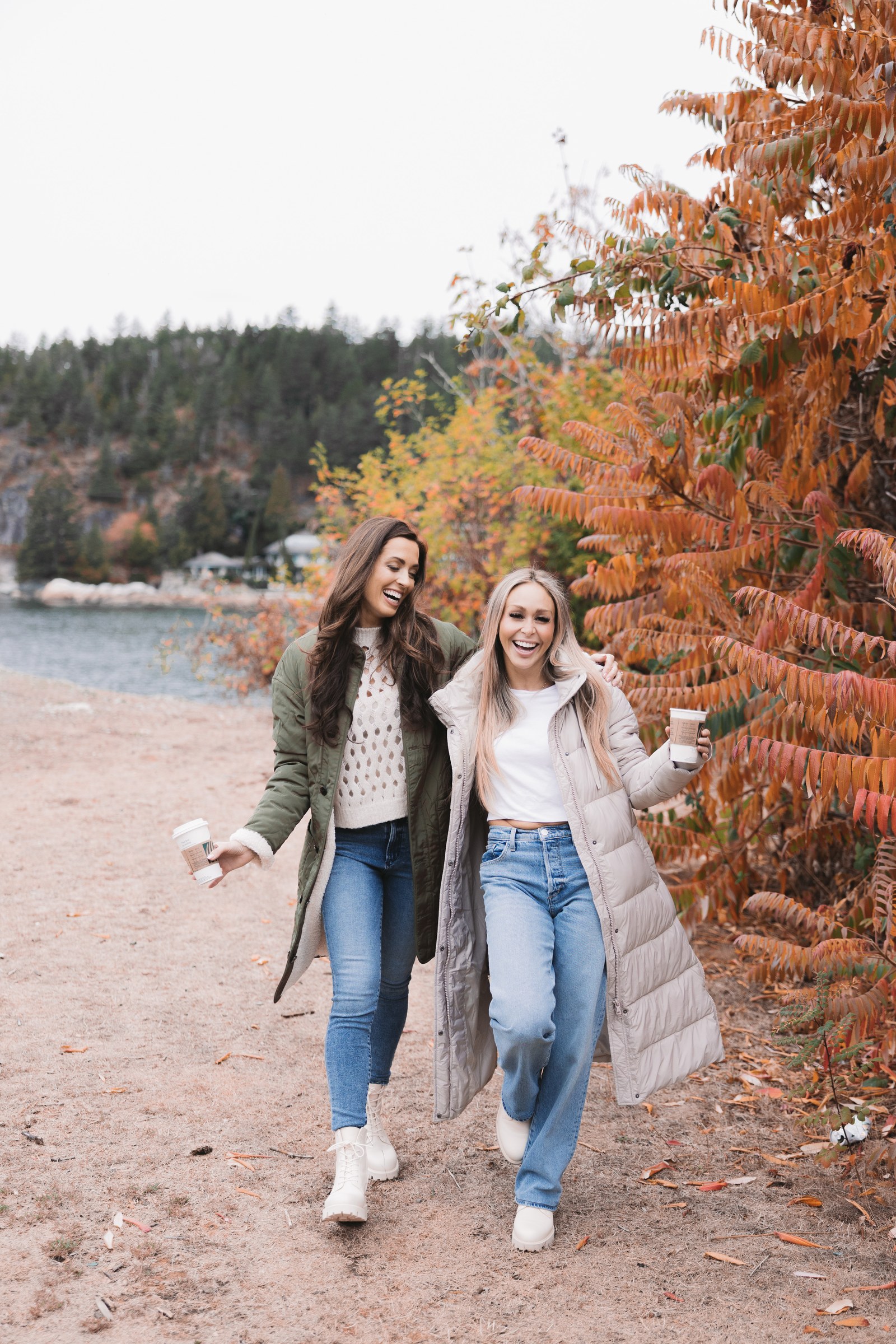 Two smiling women wearing autumn coats and jeans walking beside colourful foliage