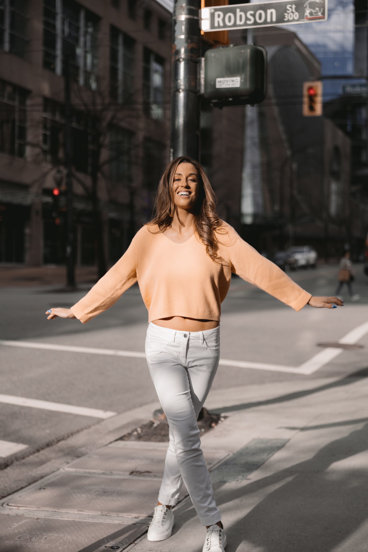 Woman wearing a peach sweater and white trousers standing at an urban crosswalk