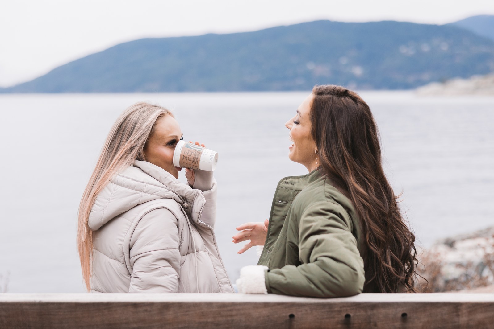 Two women in winter jackets sharing coffee on a bench beside a lake