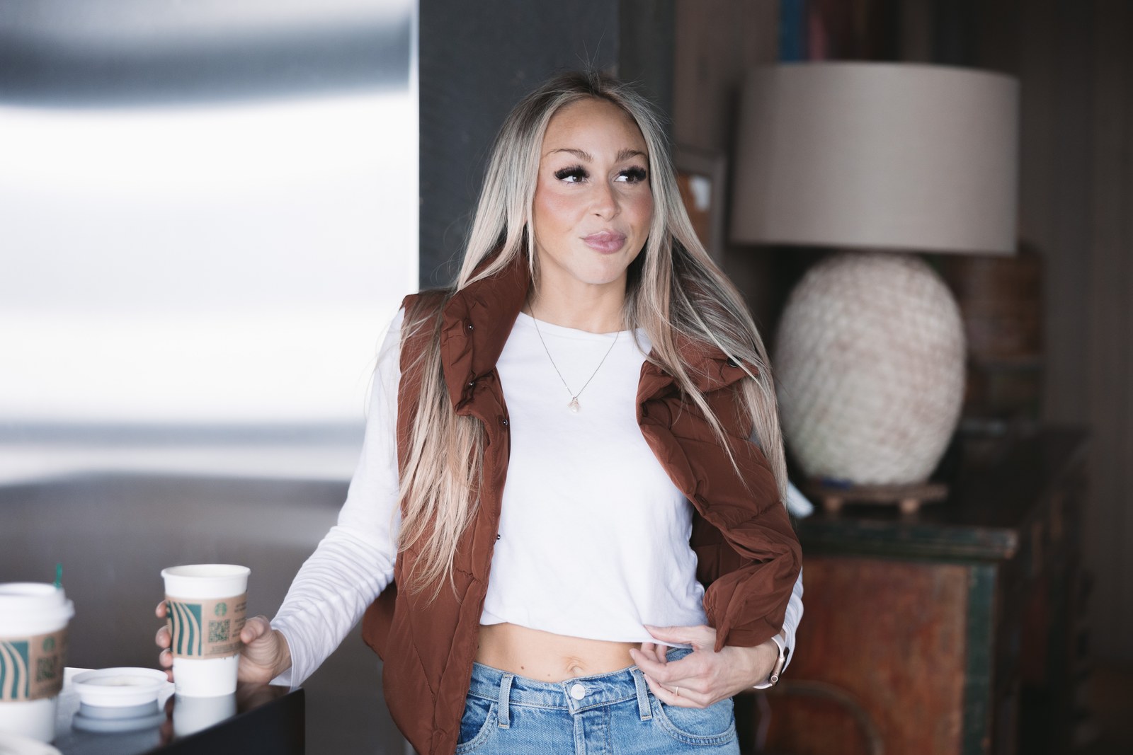Woman wearing a rust-coloured vest over a white top and jeans beside a kitchen counter