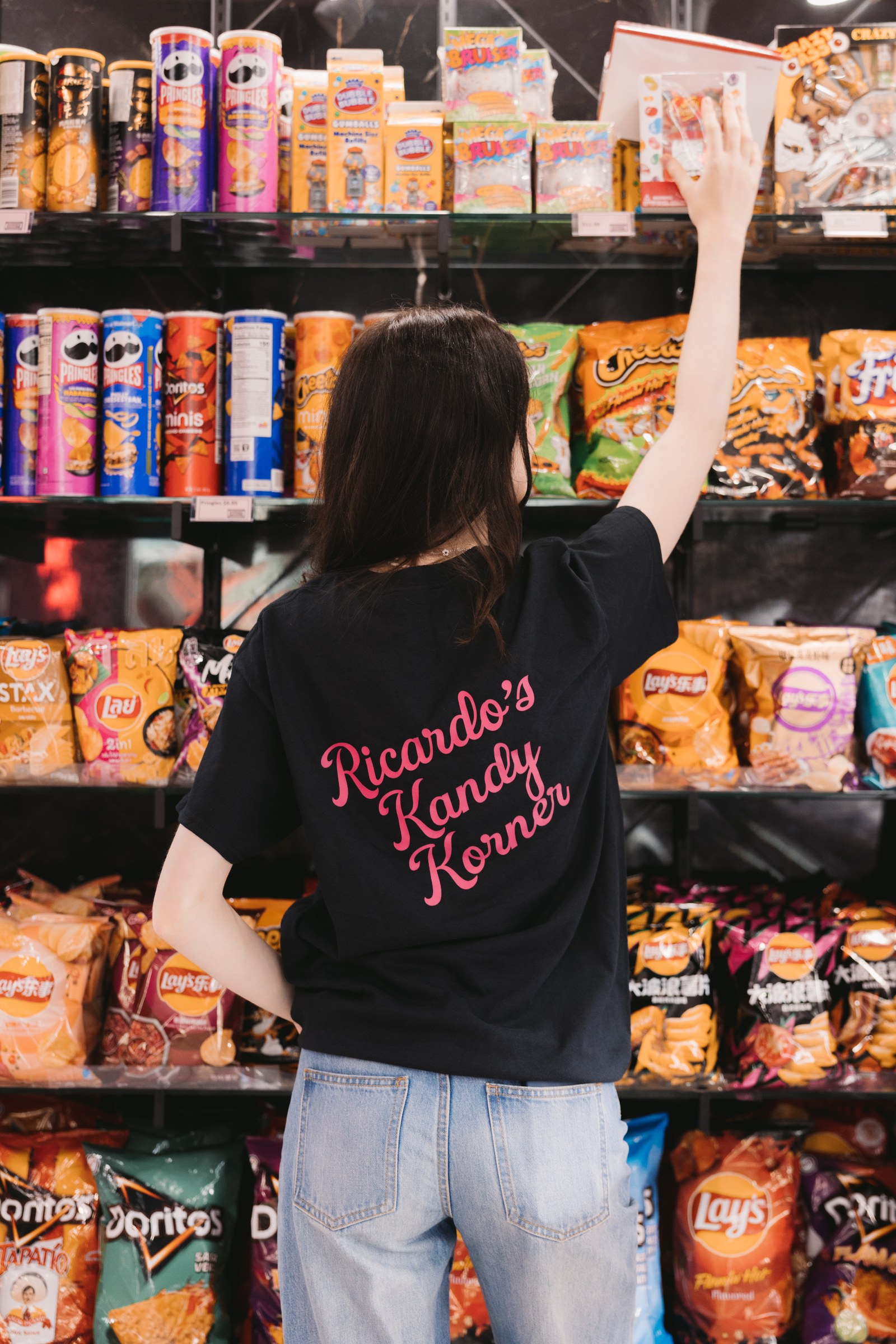 Woman wearing a black Ricardo’s Kandy Korner shirt reaching for a sweet on a shop shelf