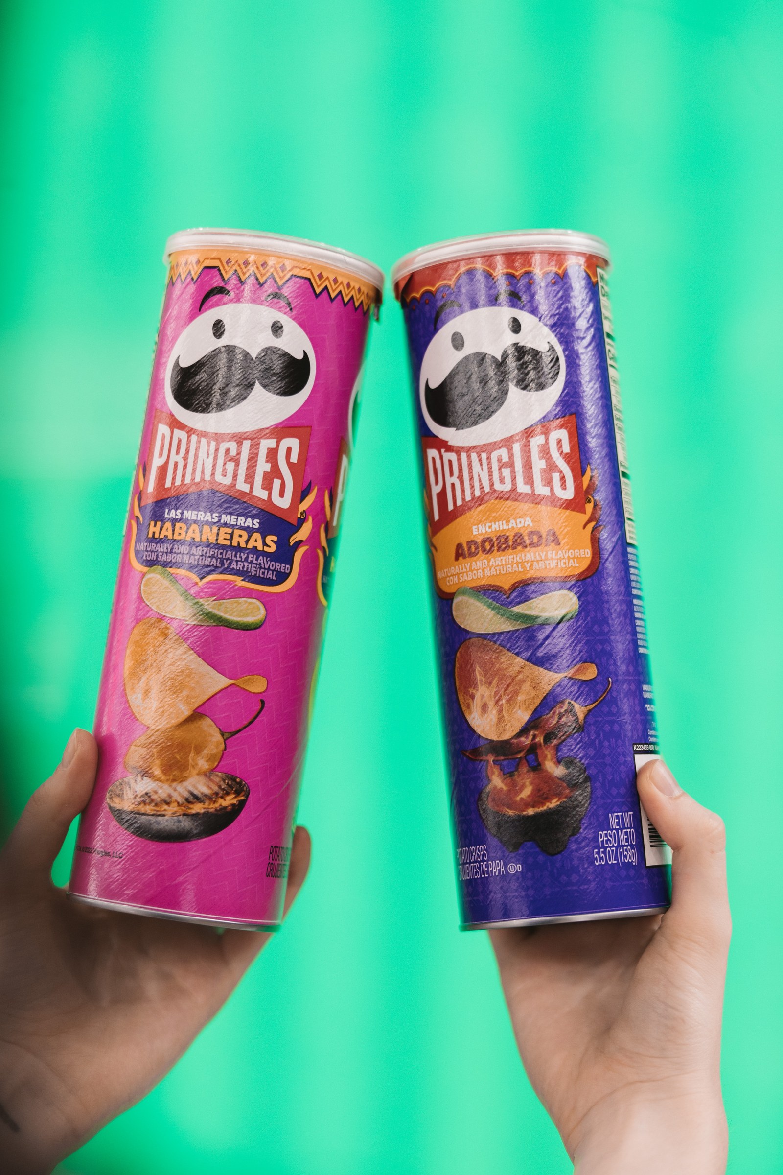 Hands holding two brightly coloured Pringles cans against a green background