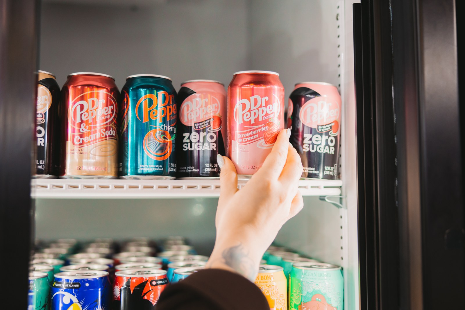 Hand selecting a pink soda can from a colourful refrigerated display
