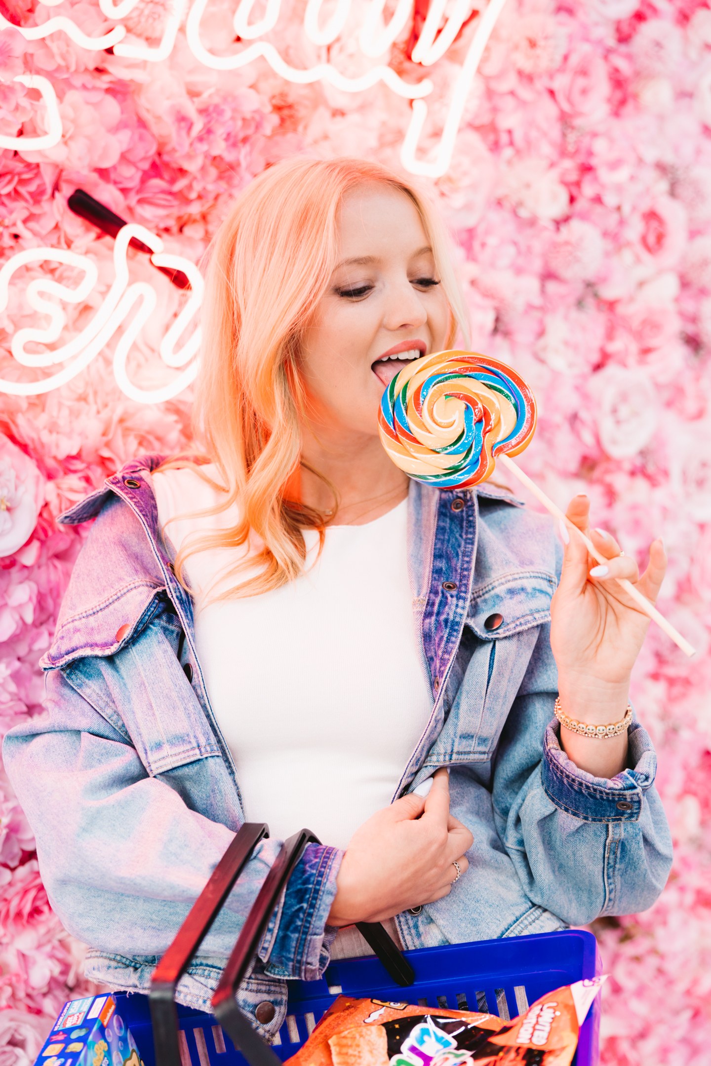 Woman in a denim jacket holding a rainbow lollipop in front of a pink flower wall