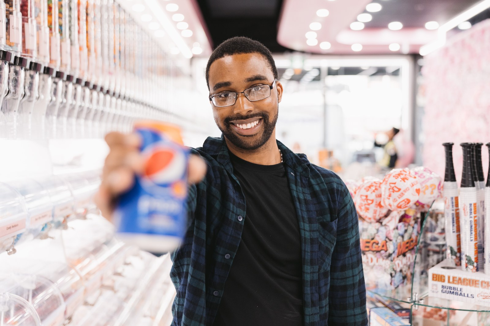 Smiling man holding a blue drink can toward the camera in a colourful candy shop