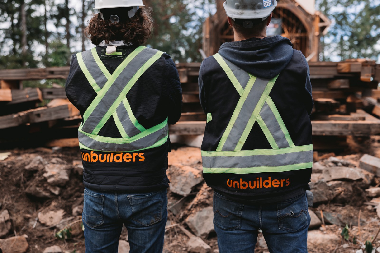 Two crew members wear Unbuilders branded safety vests in front of stacked timber