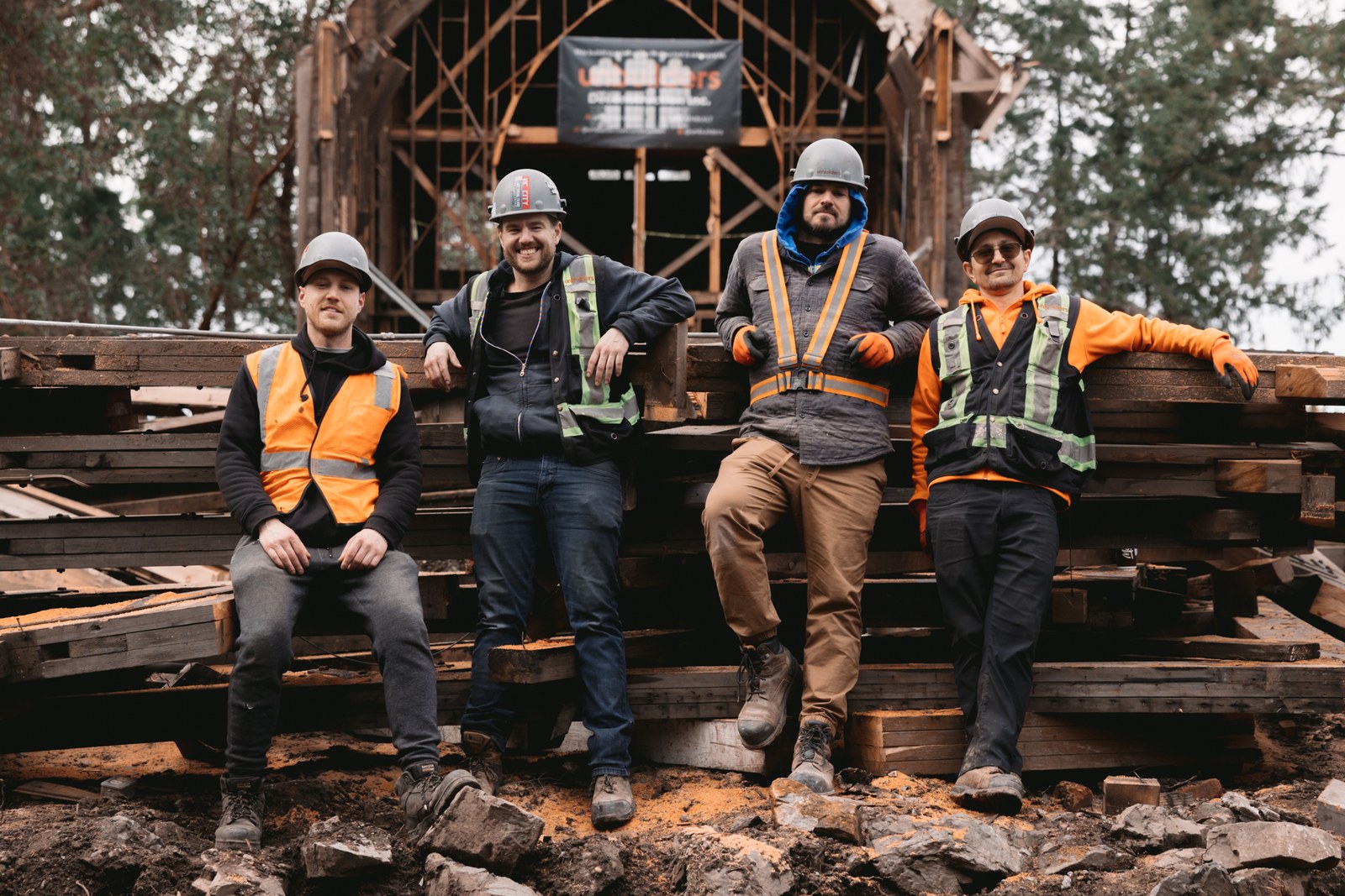 Four workers in hard hats and high-visibility vests pose beside stacked timber at a deconstruction site