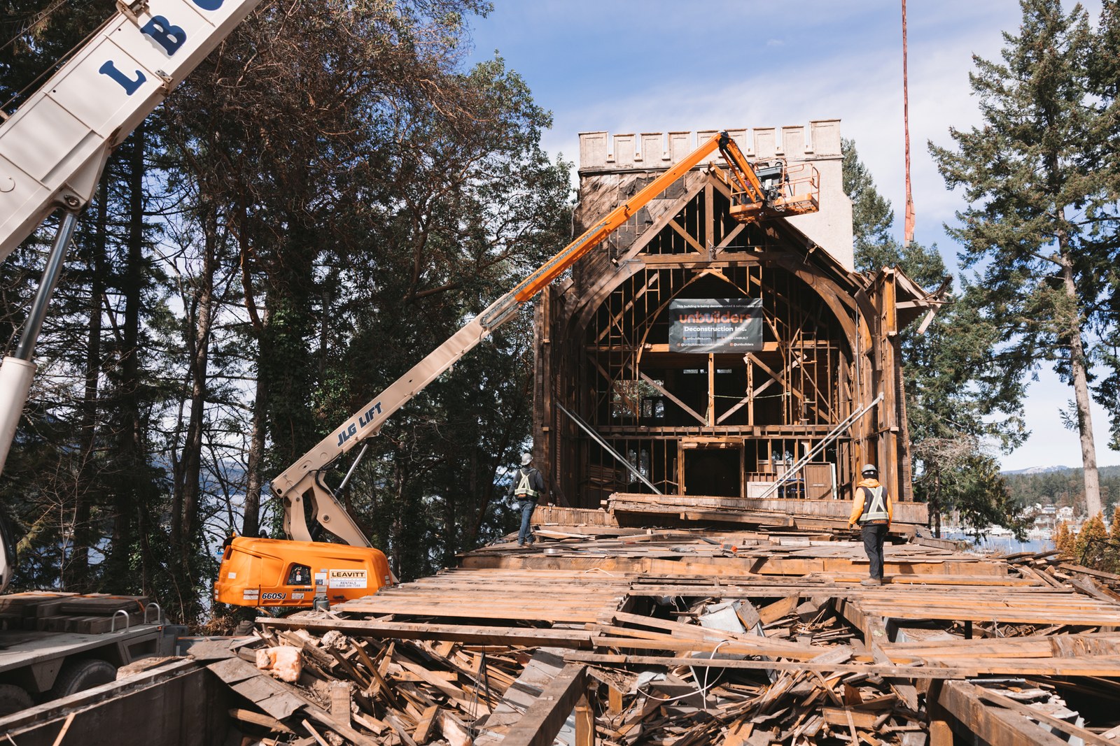 Workers and an elevated boom lift surround an exposed timber building during deconstruction