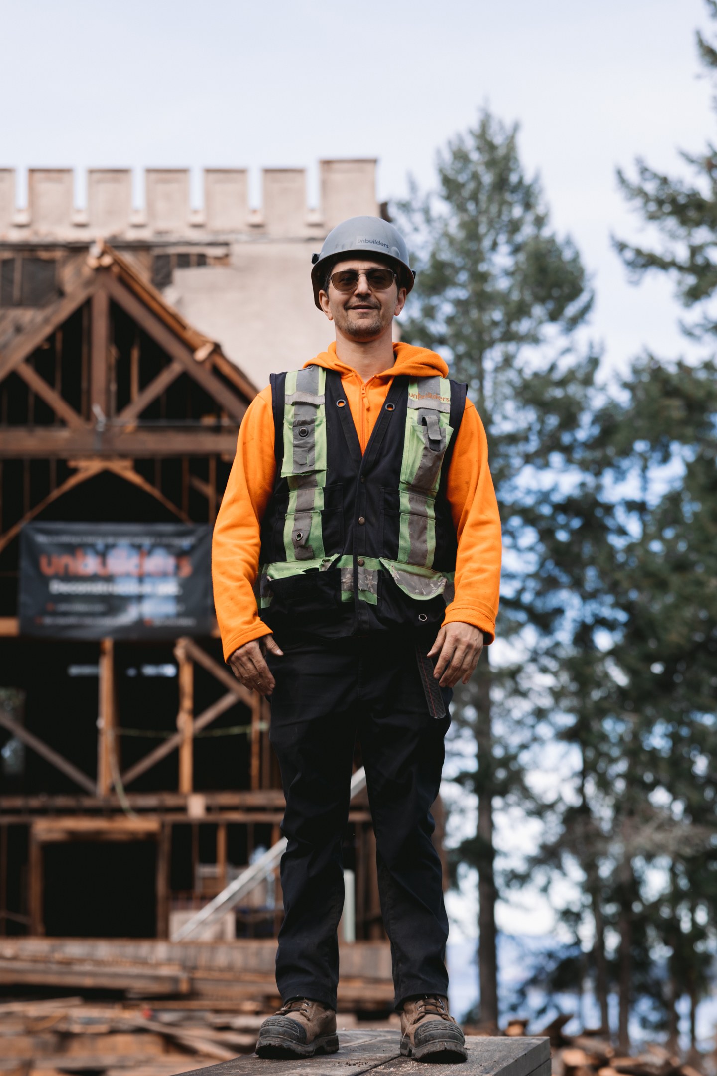 An Unbuilders crew member in an orange hoodie and hard hat stands in front of exposed timber framing