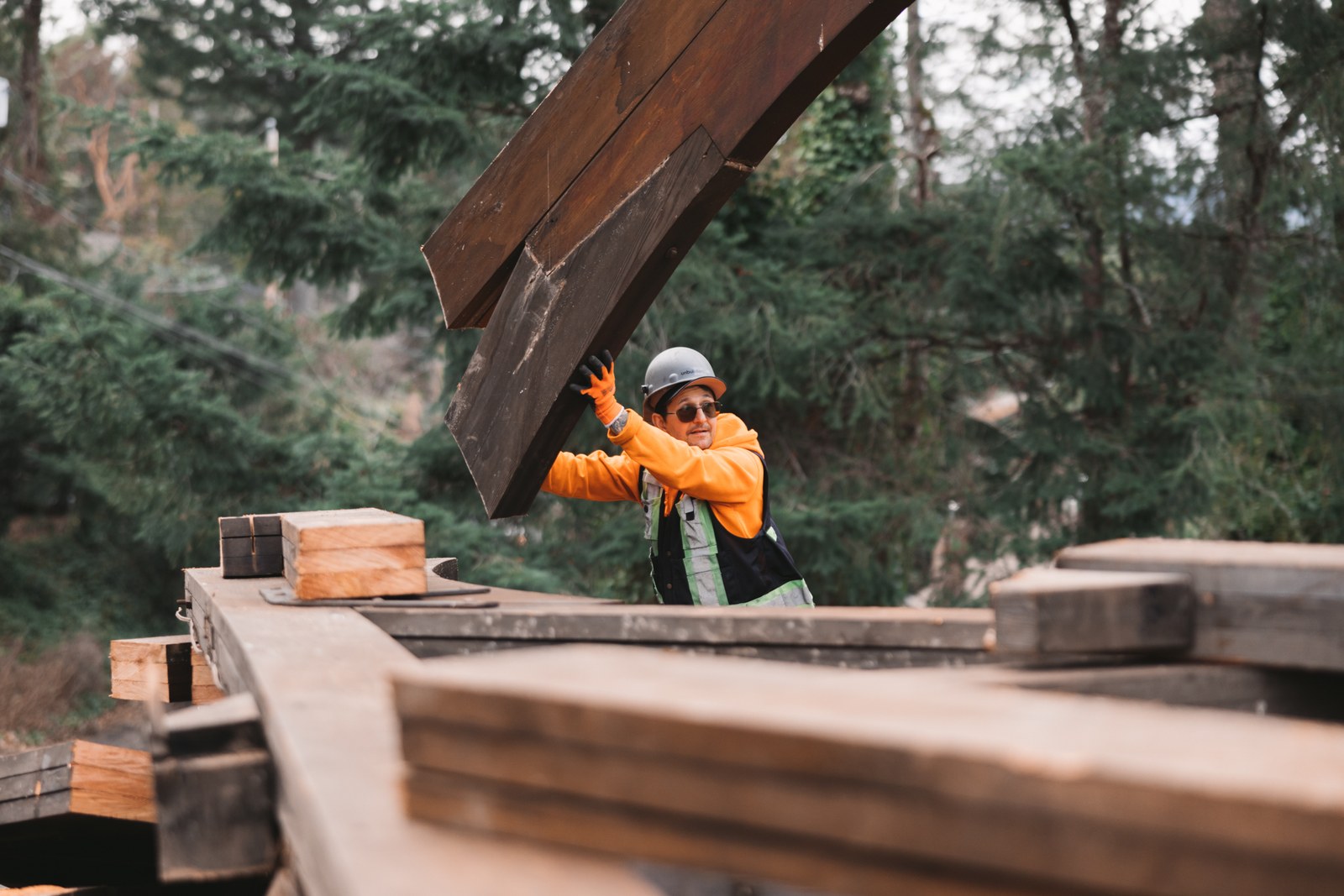 A worker in an orange hoodie and hard hat guides a large wooden beam beside stacks of timber
